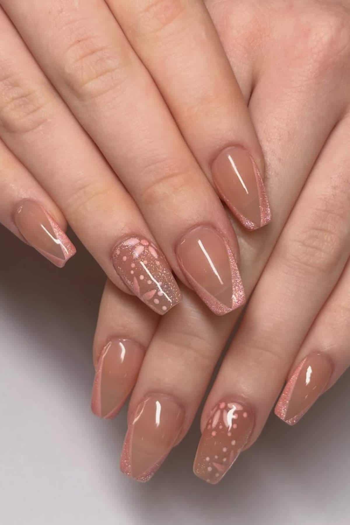 Hands with long, almond-shaped nails painted in a nude pink color with glittery tips and white dotted floral designs on two accent nails.