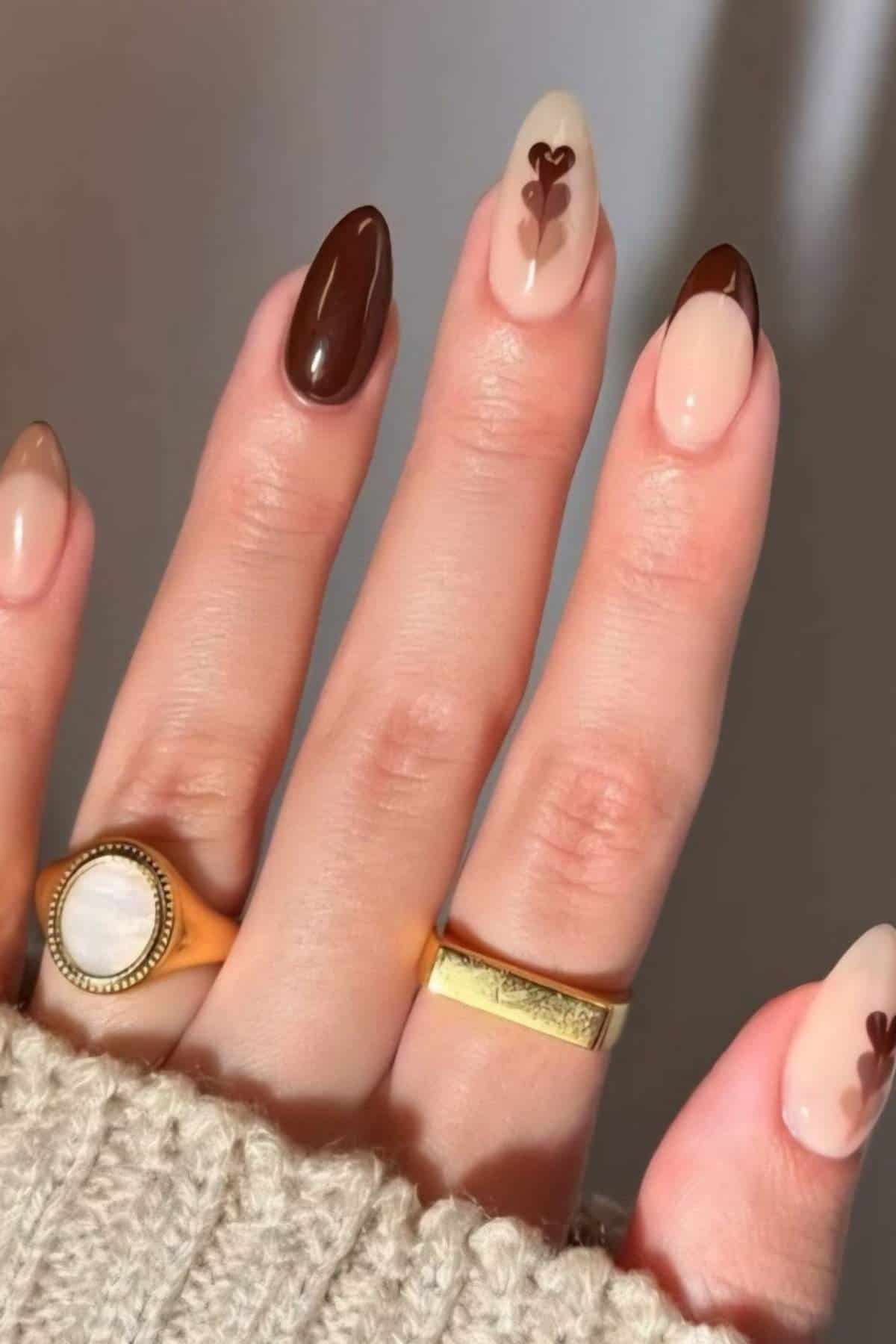 A hand with almond-shaped nails featuring a brown and nude color scheme, with two nails displaying small brown heart designs. The person wears two gold rings and a beige sweater.