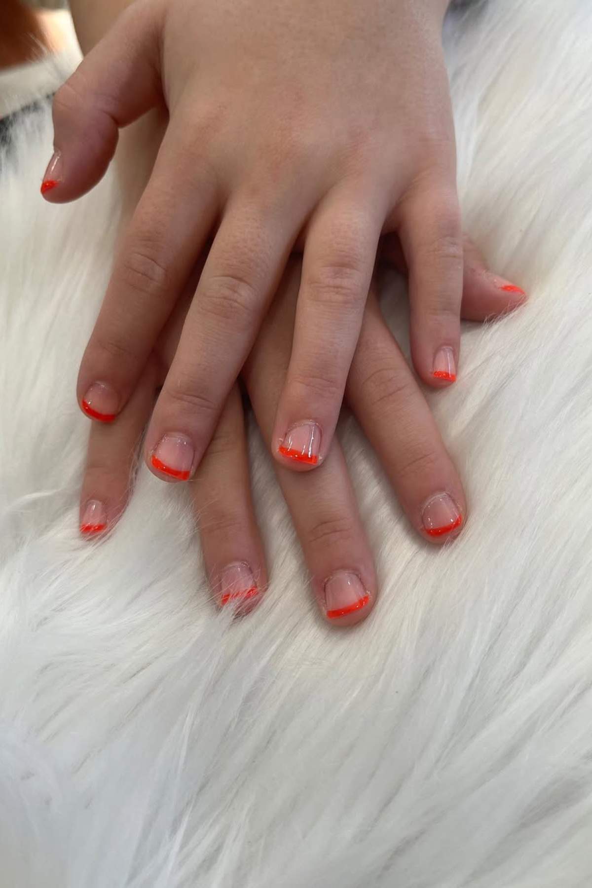 Two hands rest on a white furry surface, displaying nails with a simple red French tip manicure.
