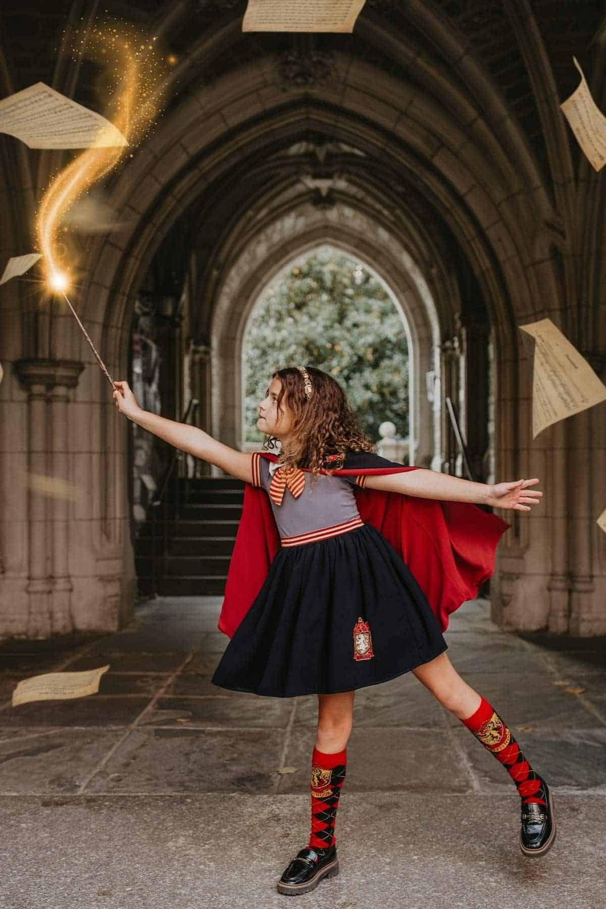 A young girl dressed as a wizard holds a wand with glowing light, surrounded by floating papers, standing under a stone archway.