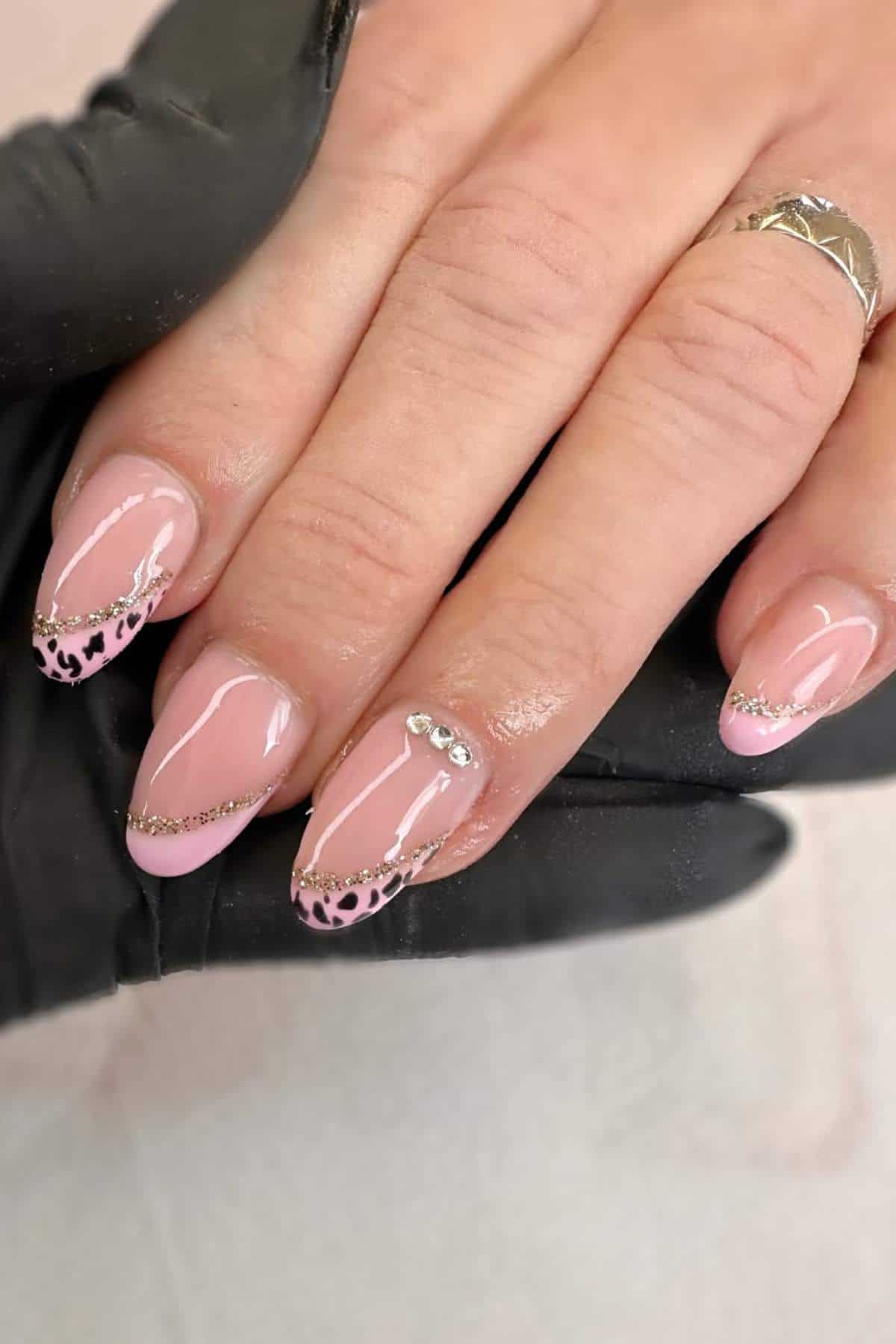 A hand with almond-shaped nails featuring a light pink base, gold glitter tips, animal print accents, and small rhinestone decorations. A black glove and silver ring are visible.