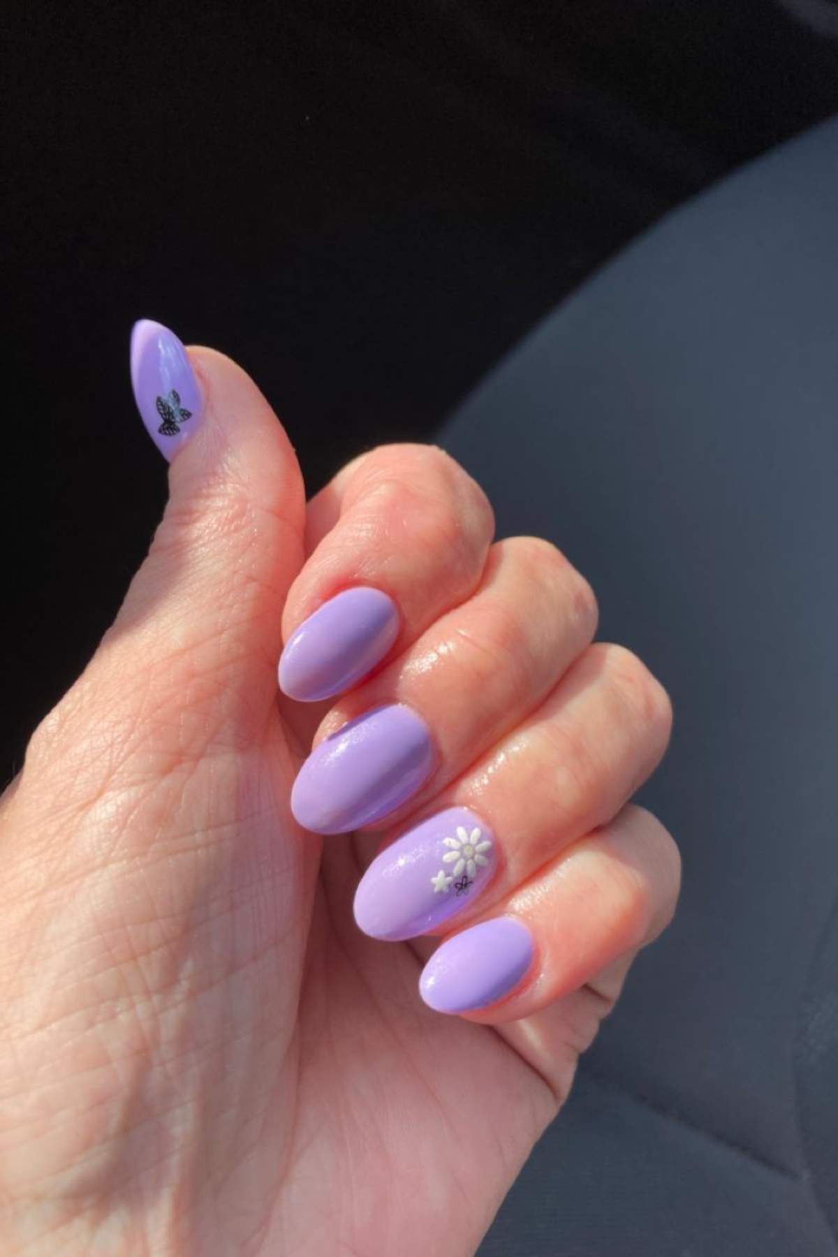 A hand with almond-shaped nails painted lavender, featuring a small butterfly design on the thumb and a white flower design on the ring finger.