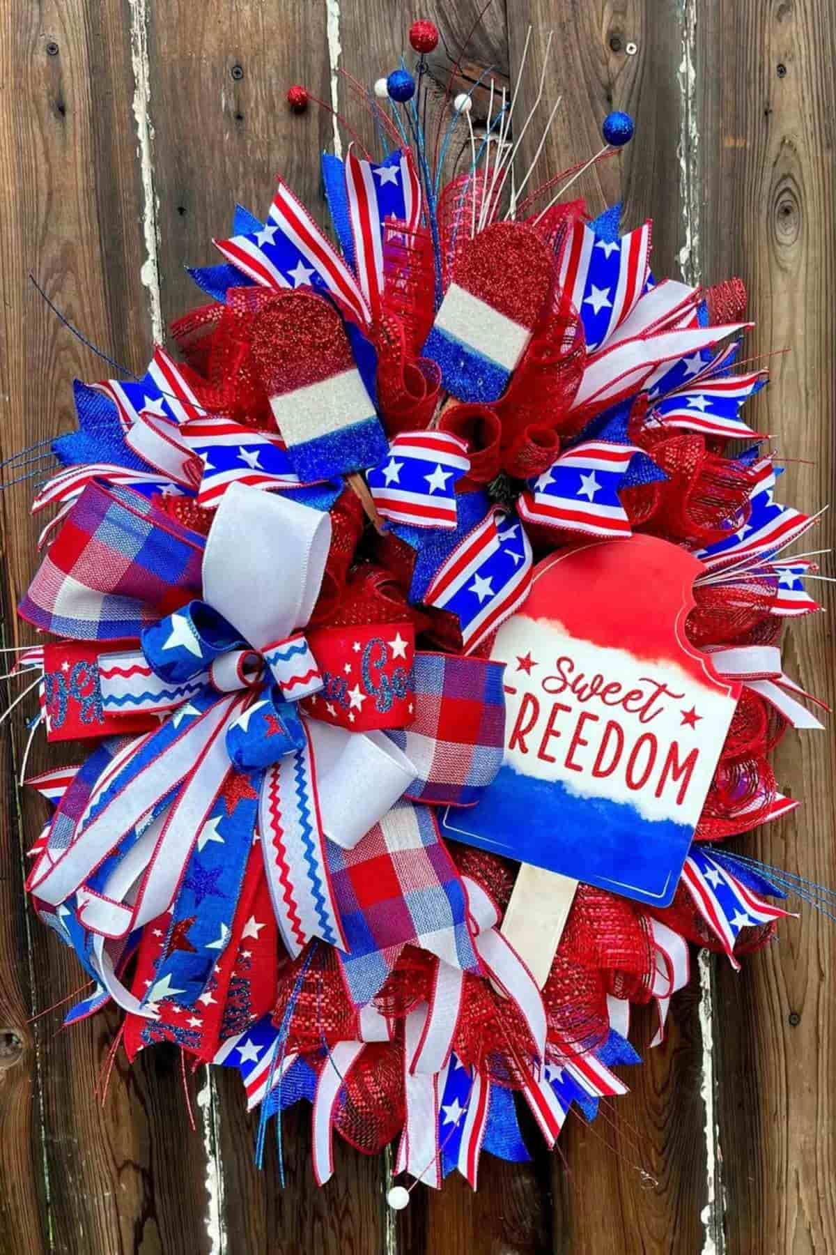Patriotic wreath with red, white, and blue ribbons, stars, and a sign reading “Sweet Freedom,” displayed on a wooden background.