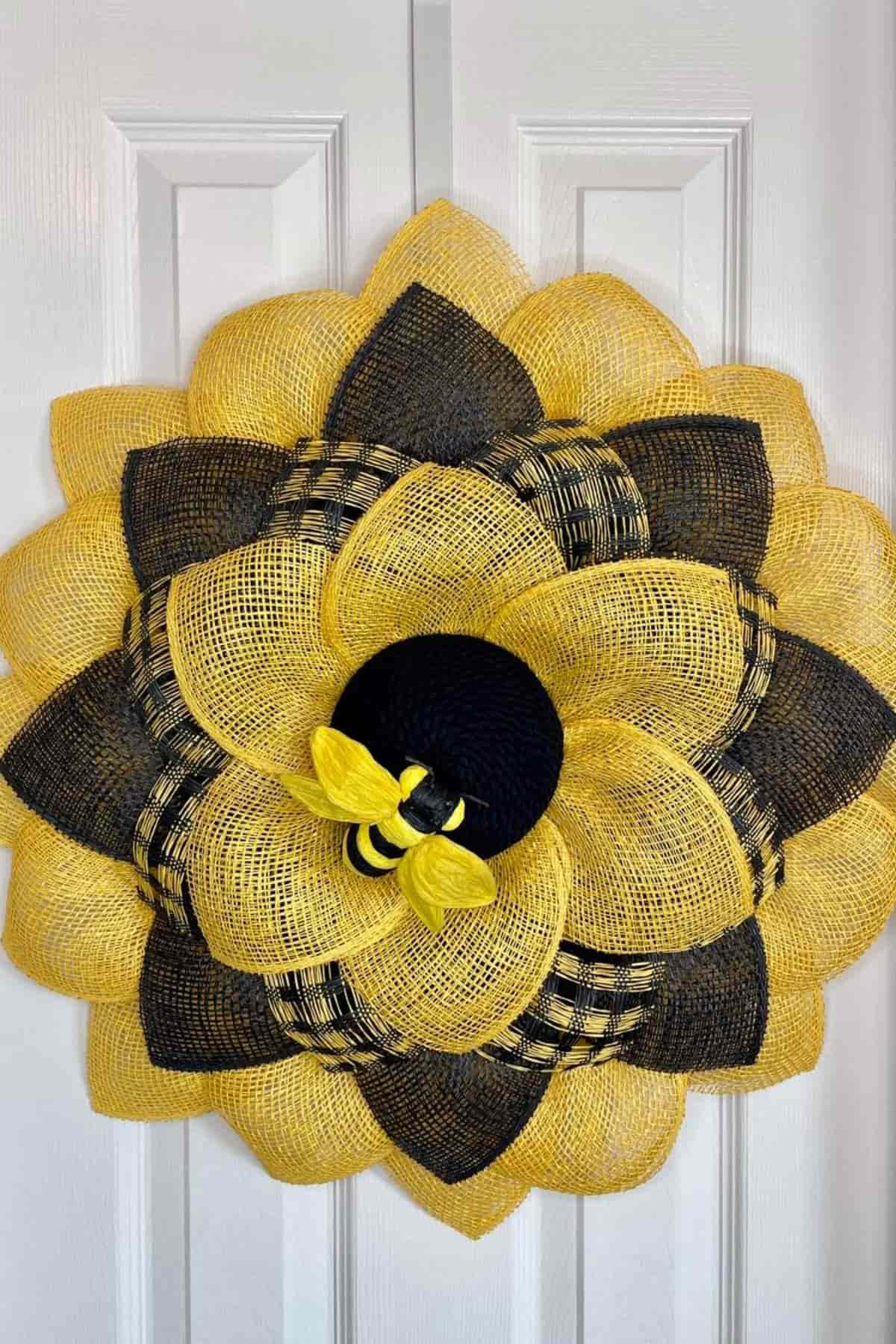 A mesh flower wreath with yellow and black petals features a decorative bee in the center, hanging on a white paneled door.