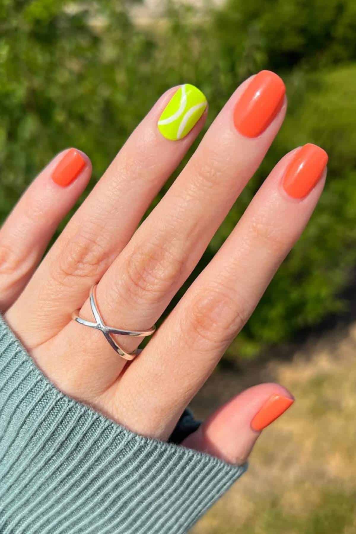 A hand with orange-painted nails and one accent nail painted neon green with a tennis ball design, wearing a silver ring, shown against a blurred outdoor background.