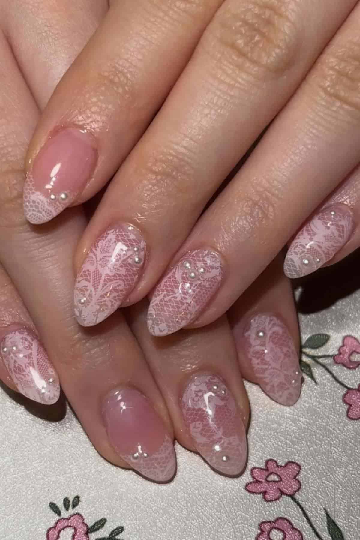 Hands with almond-shaped nails featuring pale pink lace-like designs and small pearl accents, resting on a floral-patterned surface.