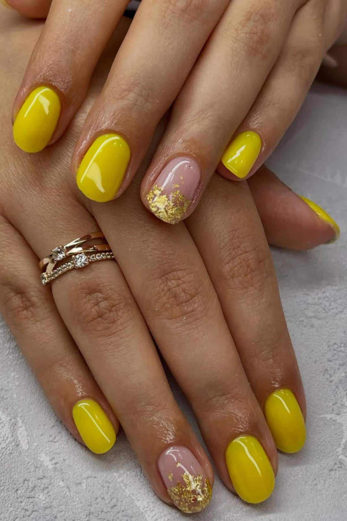 Hands with short nails painted yellow; ring finger nail is pink with gold foil decoration. Person is wearing two rings on one finger.