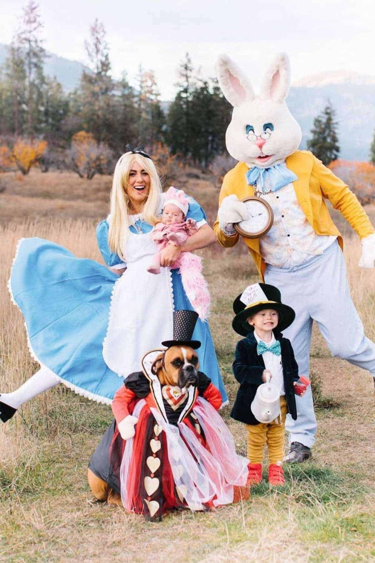 A group of people and a dog dressed in "Alice in Wonderland" costumes pose outdoors in a grassy field with trees and mountains in the background.