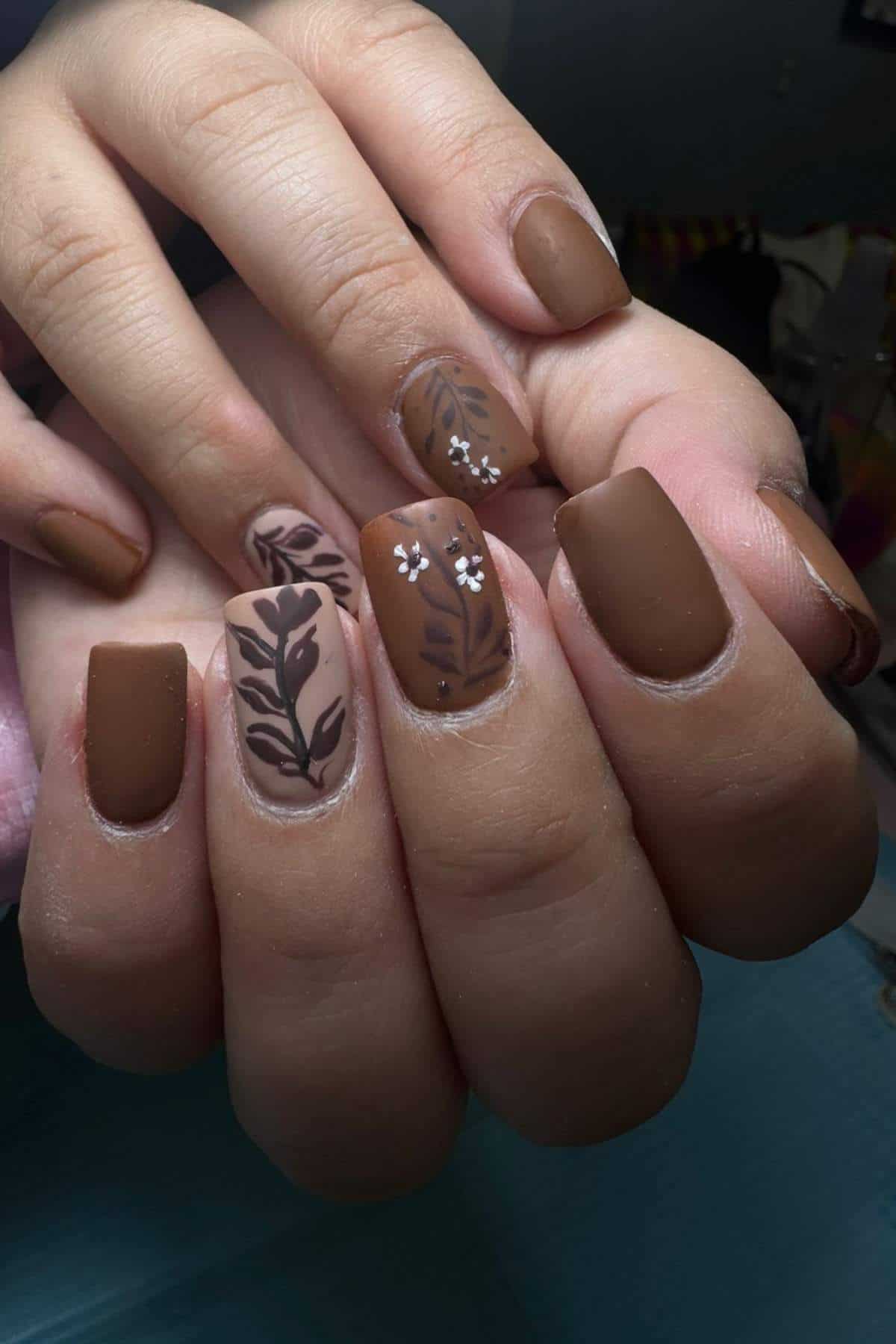 Close-up of hands with matte brown nails featuring leaf and small white flower designs on an accent nail.