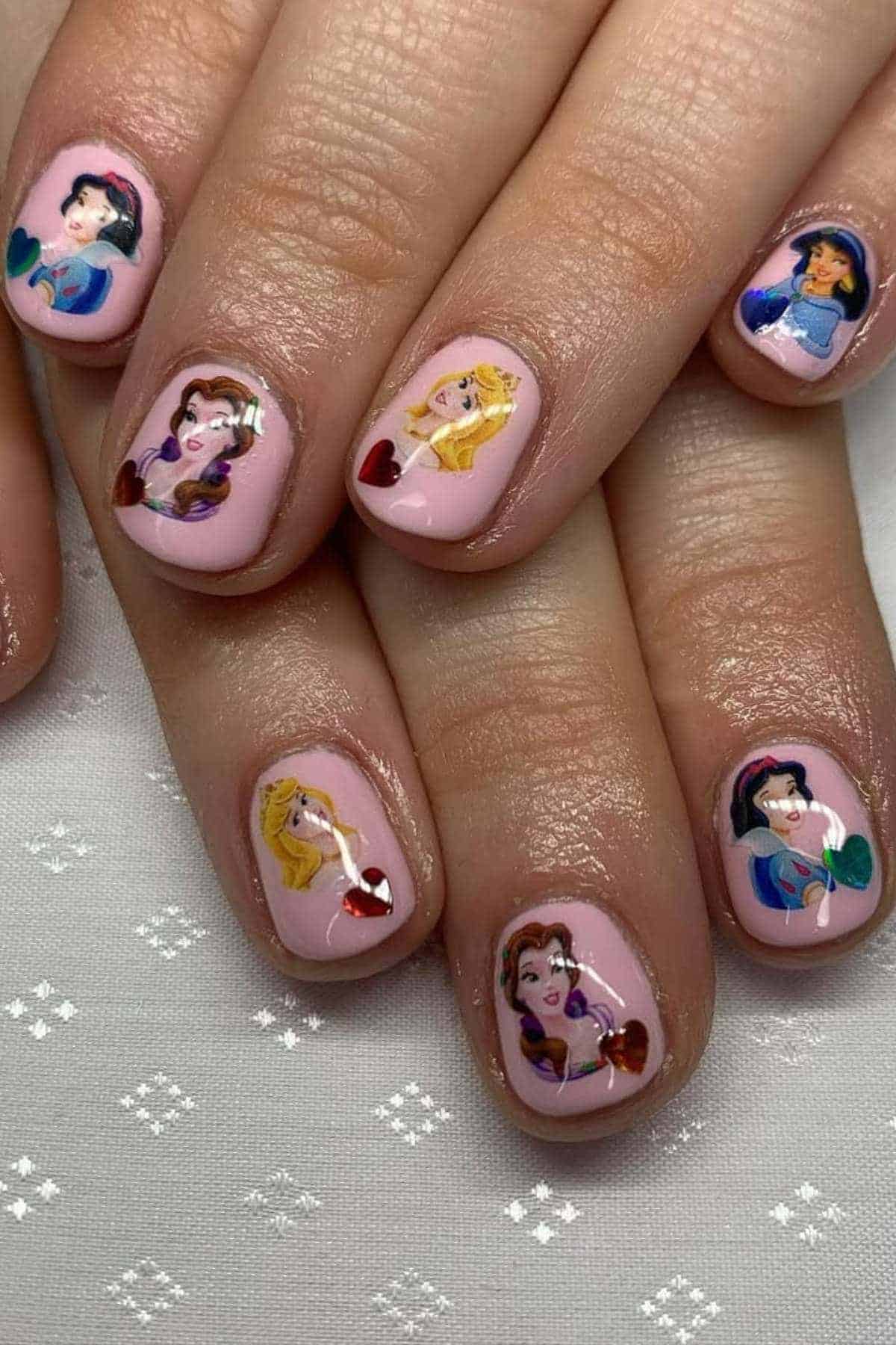 Close-up of hands with pink nail polish featuring detailed illustrations of animated princess characters on each fingernail.