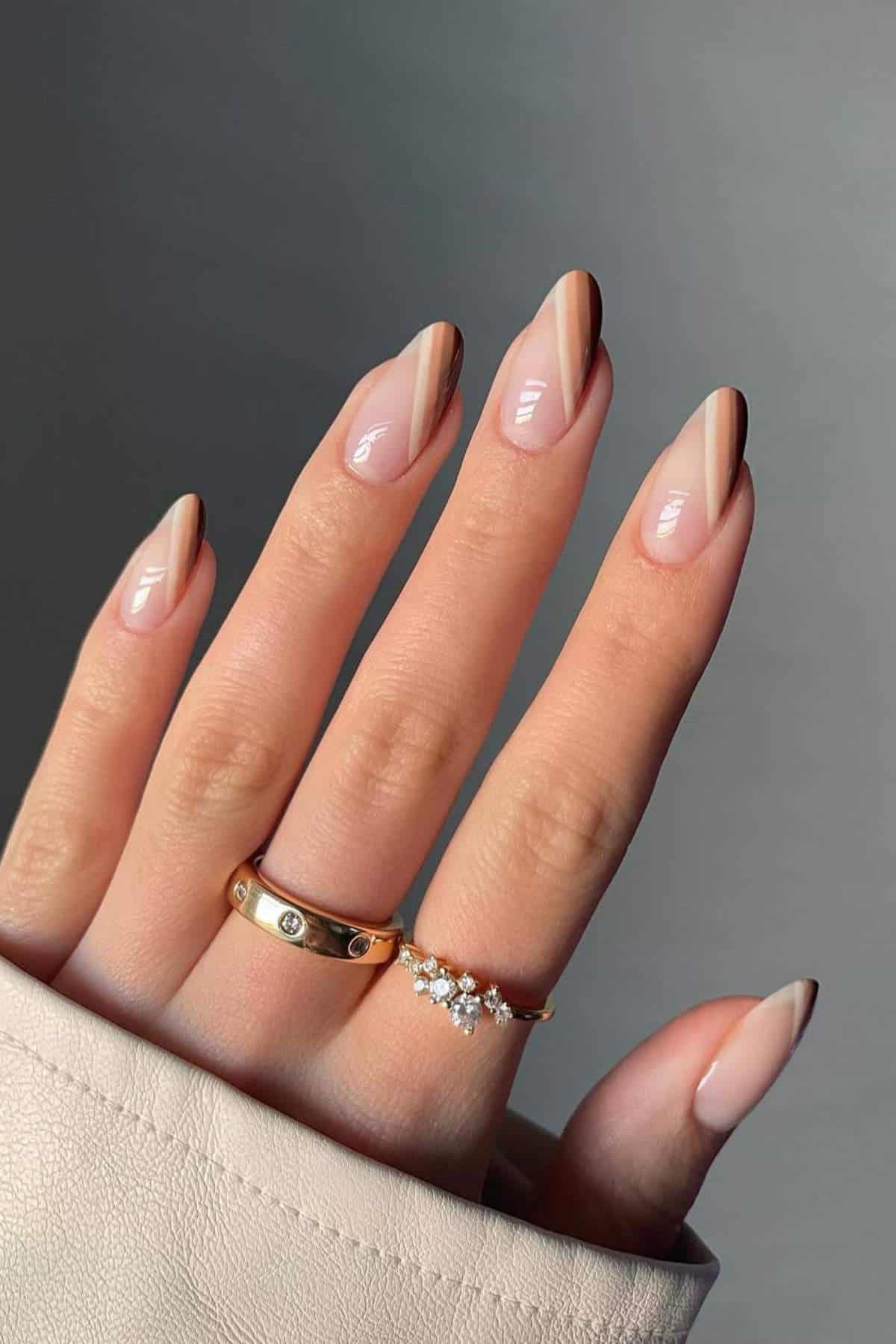 A hand with almond-shaped nails featuring a minimalist brown and nude striped design, wearing two gold rings, against a neutral background.