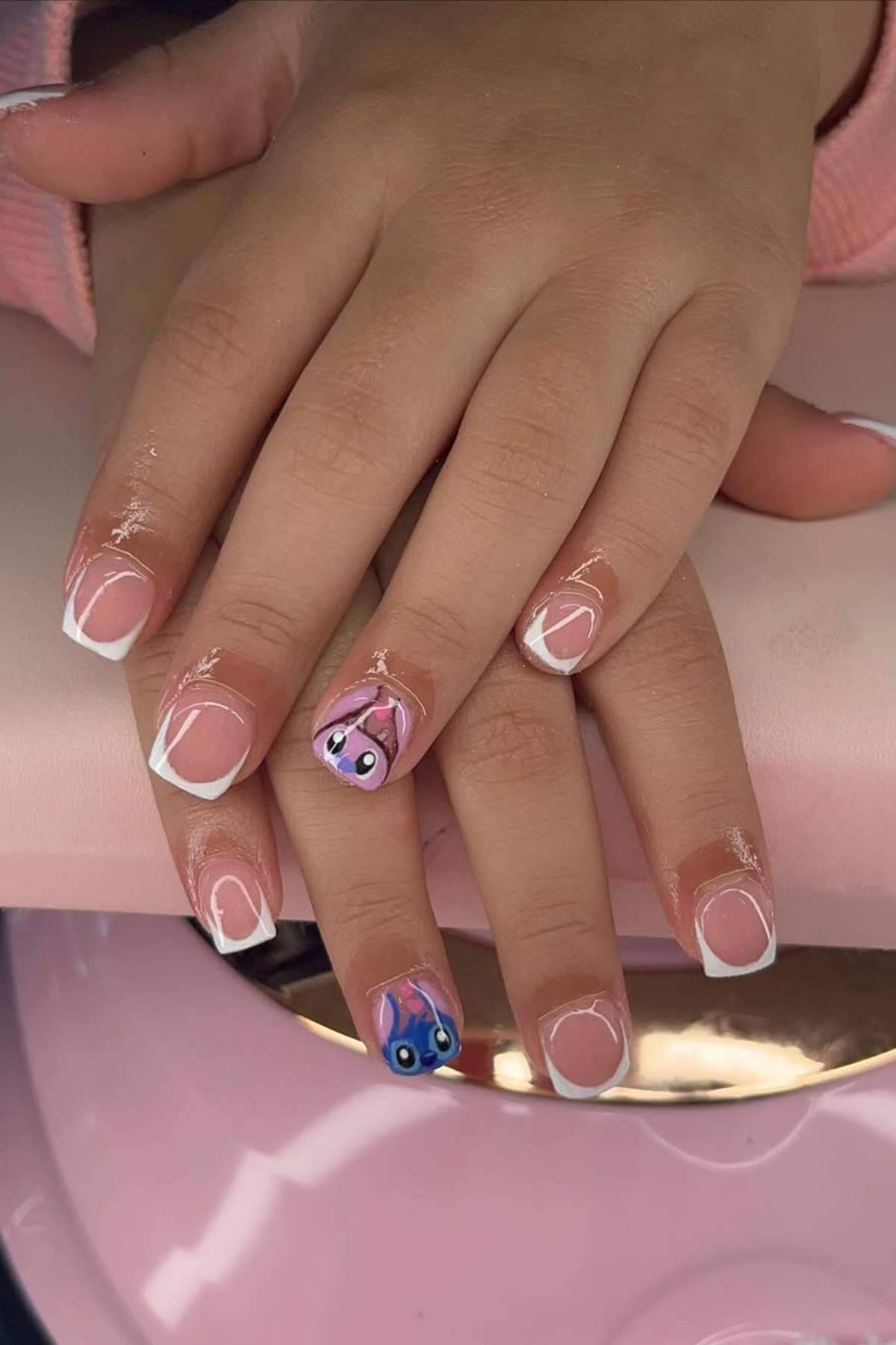 Two hands with short nails feature French tips and cartoon character nail art on the ring fingers, one pink and one blue, against a light pink background.