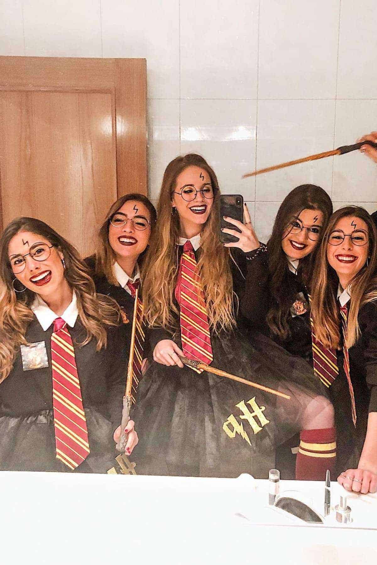 Five people dressed in Harry Potter costumes, wearing glasses and Gryffindor ties, pose and smile for a selfie in front of a bathroom mirror holding wands.