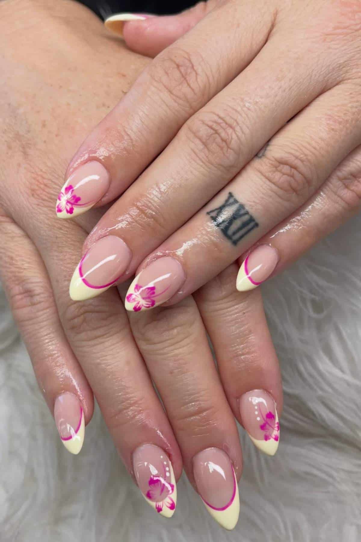 Hands with almond-shaped nails featuring a yellow French tip and pink flower nail art; a Roman numeral tattoo is visible on one finger.