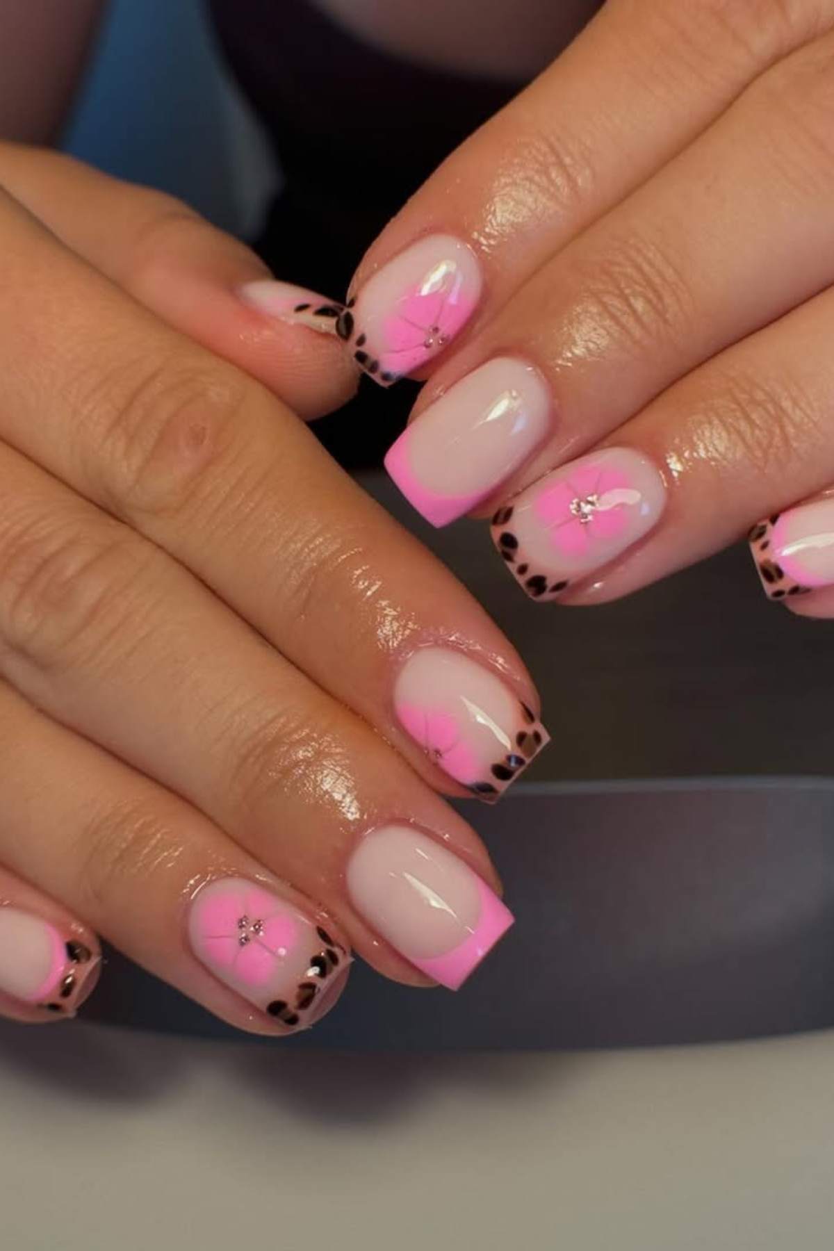 Hands with manicured nails featuring pink French tips, black leopard print on the edges, and small rhinestone decorations on a pale pink base.