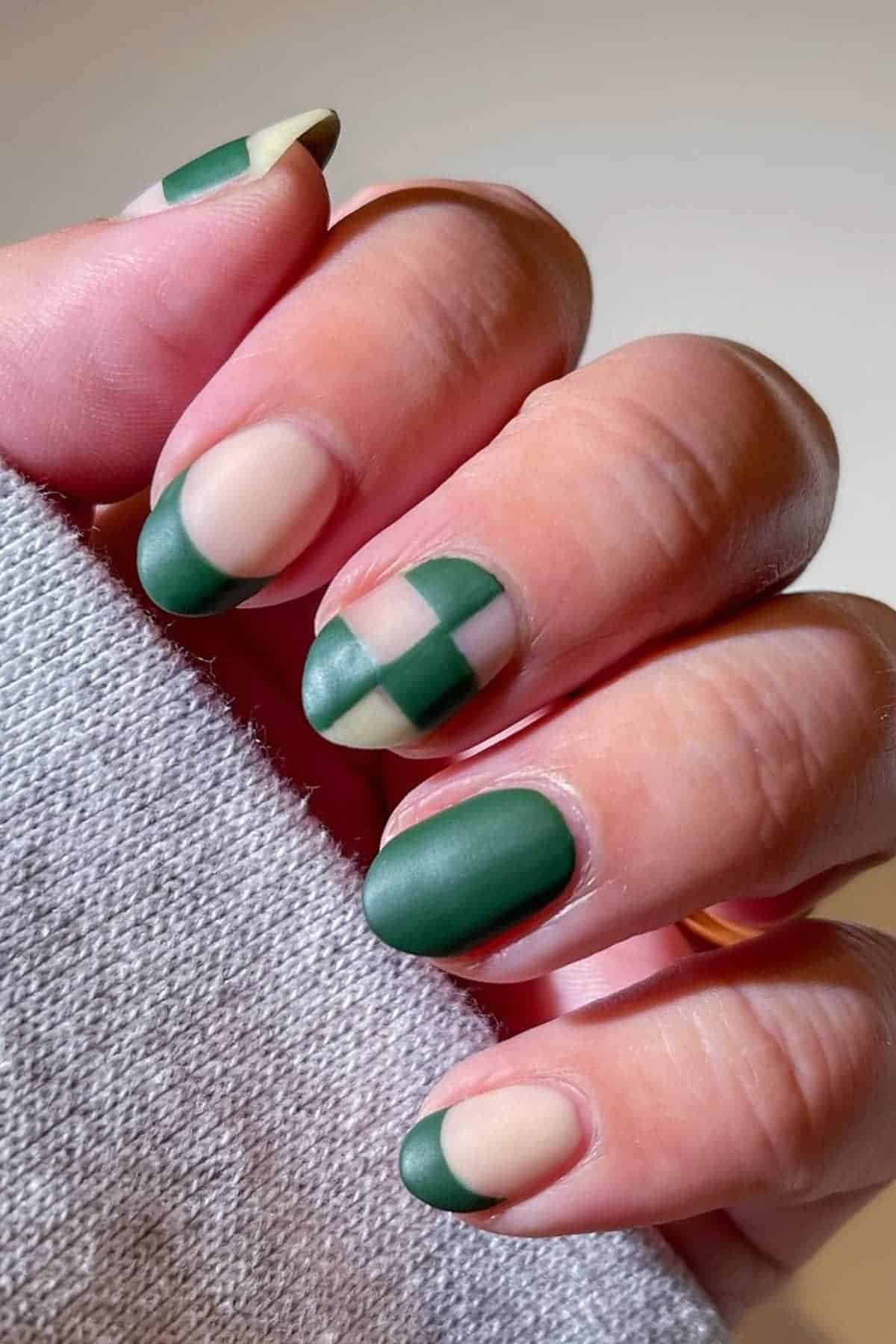 A hand with short almond-shaped nails featuring matte green and beige geometric nail art designs, including color blocking and checkerboard patterns.