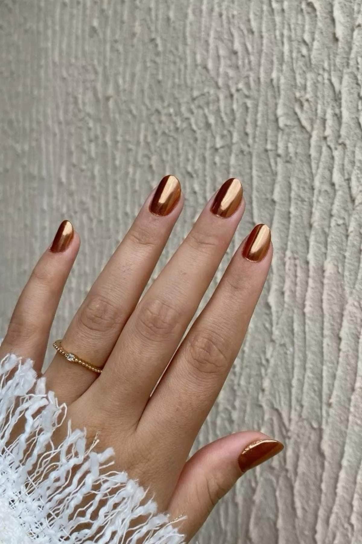 A hand with a gold ring and metallic bronze and gold nail polish is held against a textured beige wall.