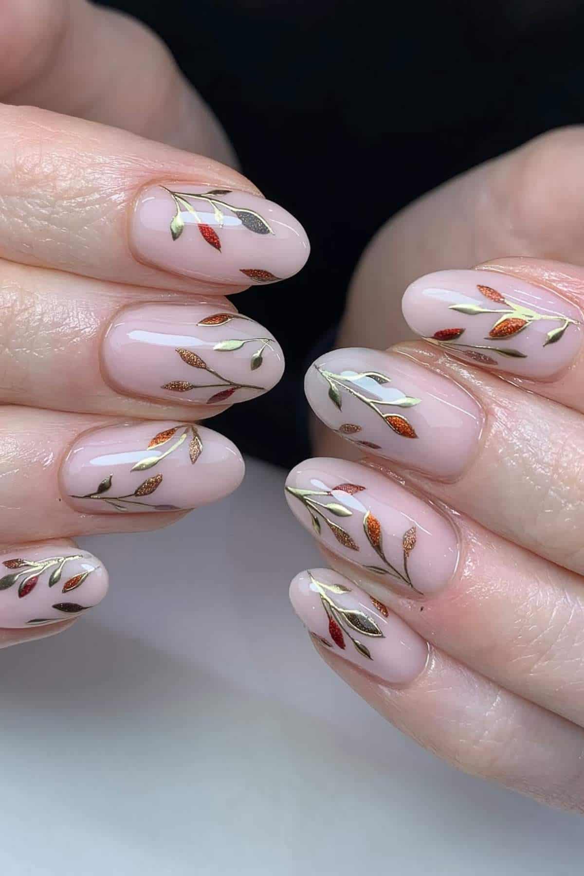 Close-up of hands with almond-shaped nails painted pale pink and decorated with metallic leaf designs in gold, bronze, and copper tones.