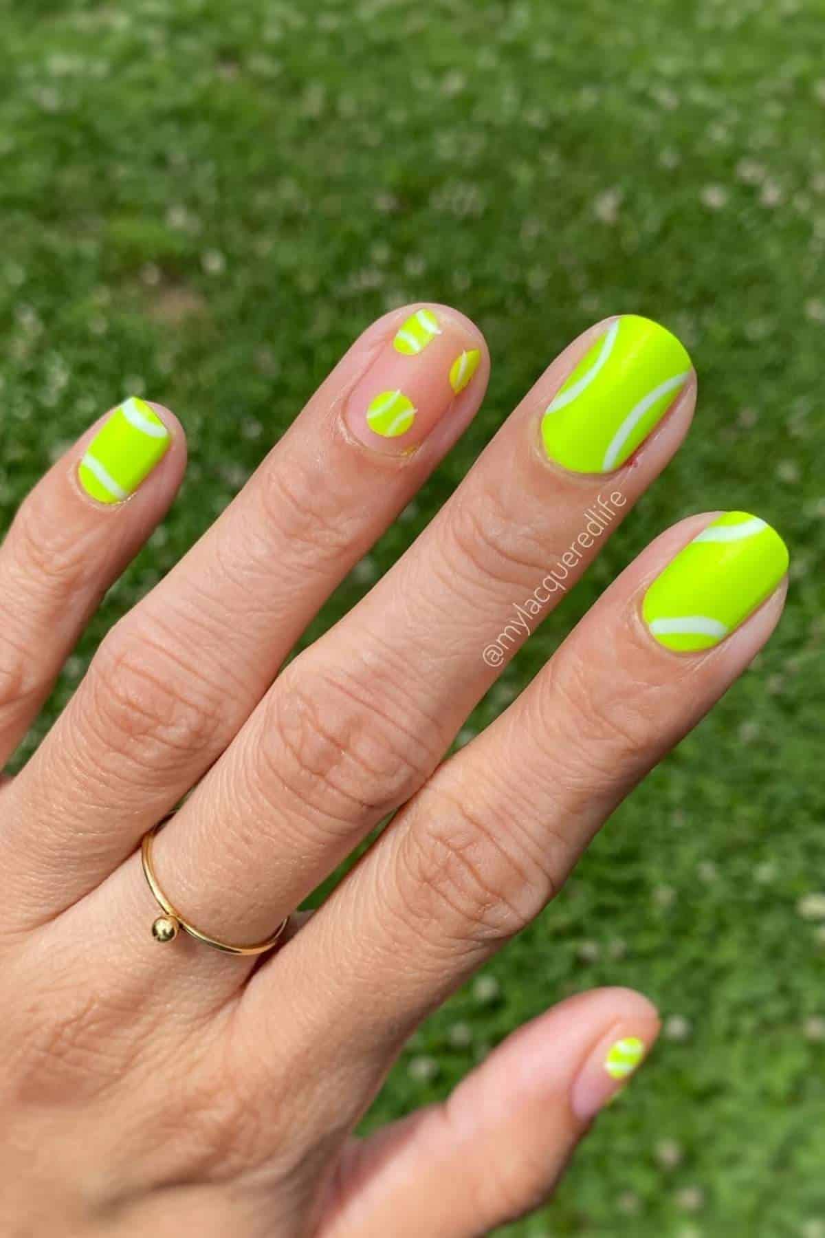 A hand with short nails painted in neon yellow with abstract clear sections, set against a blurred green grass background.
