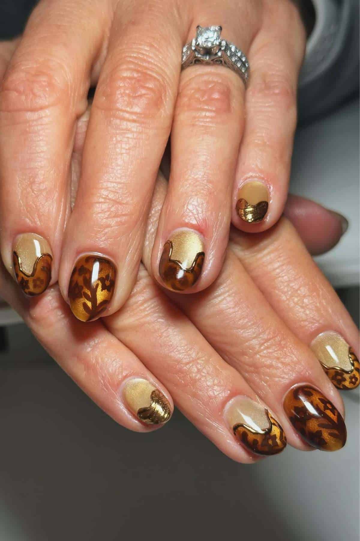Hands with manicured nails featuring a brown, gold, and animal print design; person is wearing a diamond ring on their ring finger.