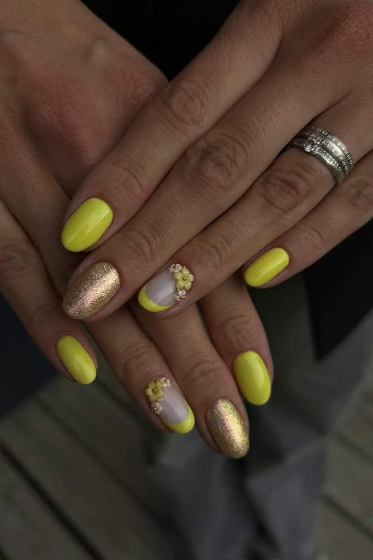Hands with yellow, gold glitter, and clear nail polish designs, featuring 3D floral accents on two nails. Two rings are worn on the ring finger.