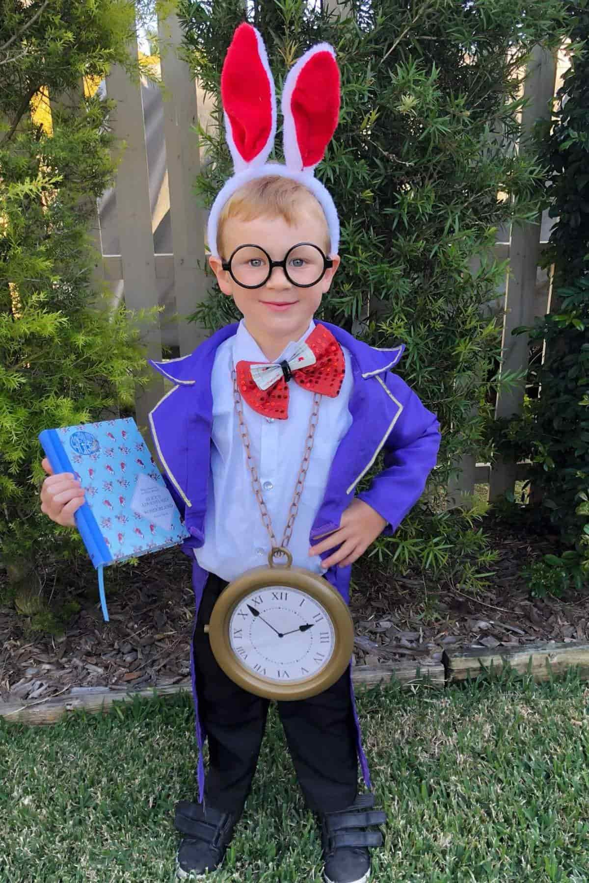 Child in a purple coat, red bow tie, and rabbit ears headband, holding a book and wearing a large clock necklace, standing on grass in front of bushes.