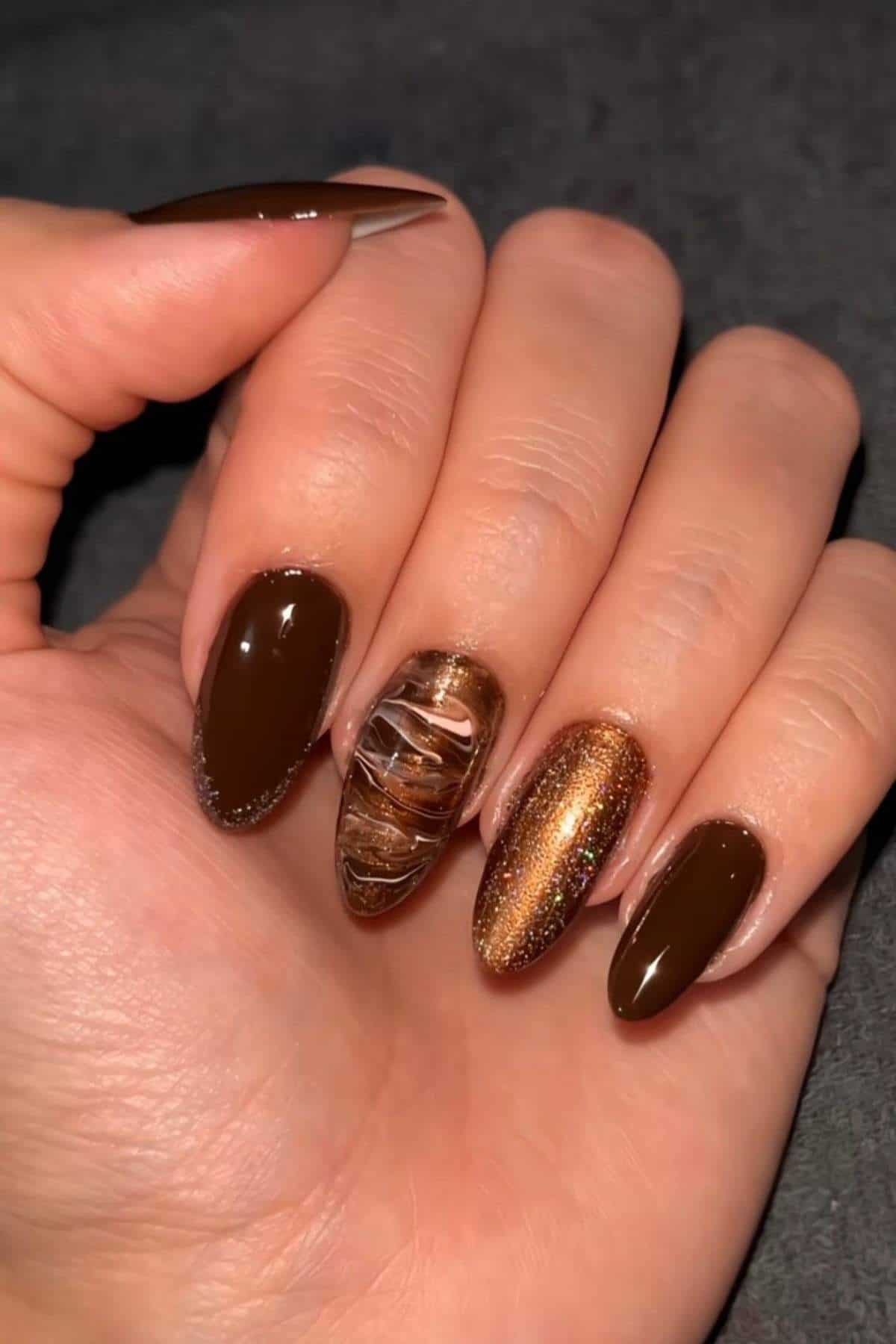 A hand with long, almond-shaped nails painted in glossy brown shades, with one nail featuring a gold glitter design and another with a marbled pattern.