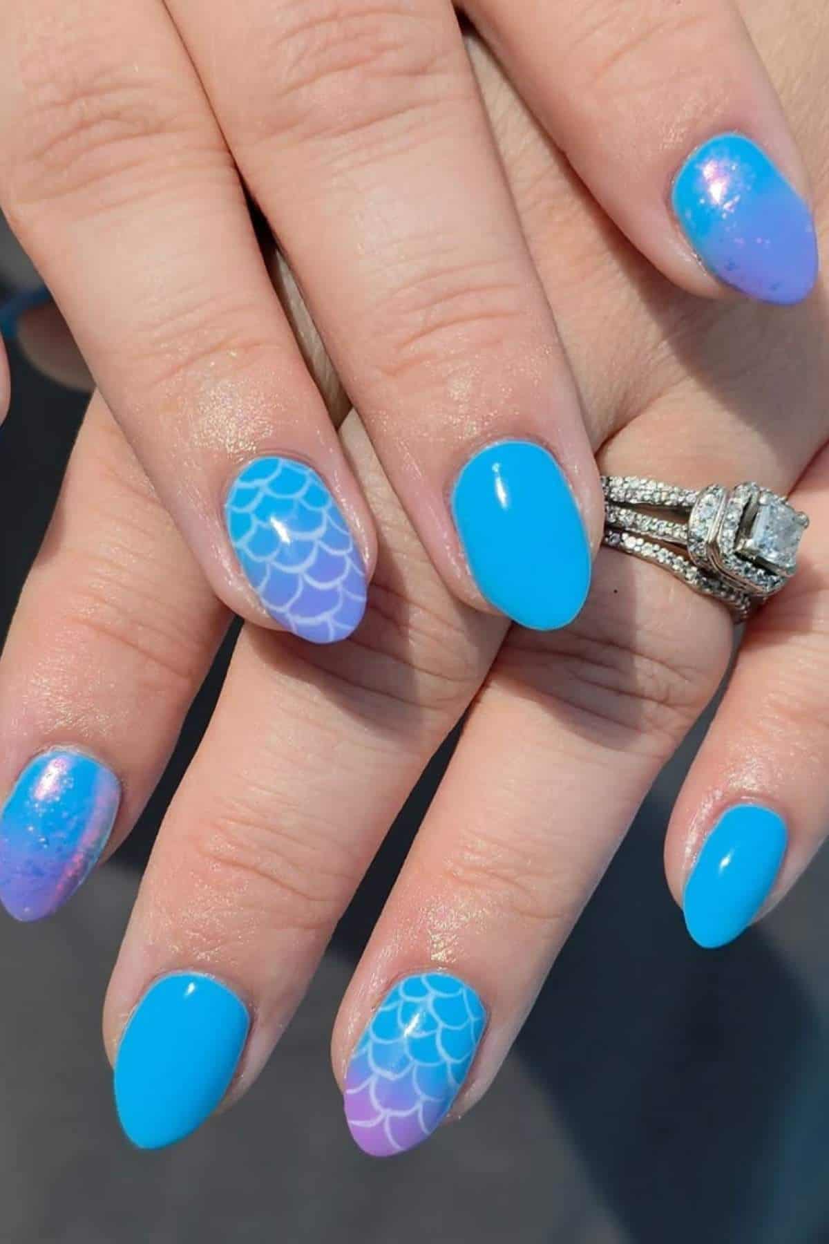 Hands with bright blue and purple manicure; some nails have white mermaid scale designs, others are solid blue or have a subtle shimmer. A diamond ring is worn on one finger.