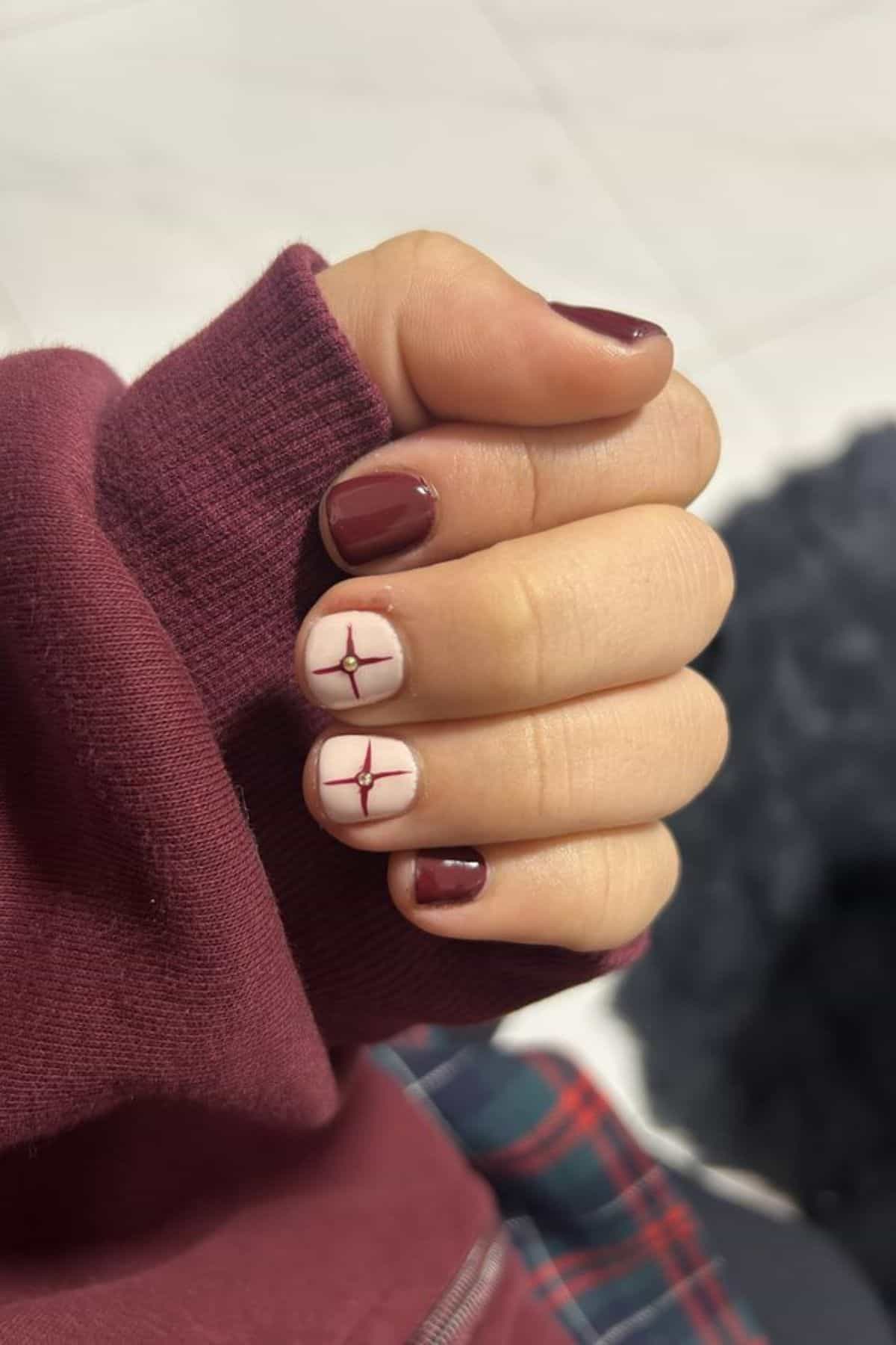 A hand with short nails painted maroon, except for two nails with a beige base and maroon geometric line designs. The person is wearing a maroon sleeve.