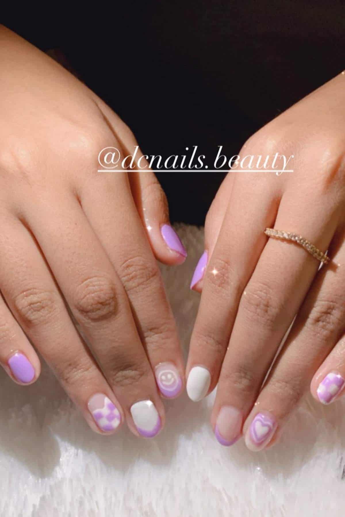 Hands with manicured nails featuring purple, pink, and white designs, including hearts, smiley faces, and checker patterns on a soft surface.