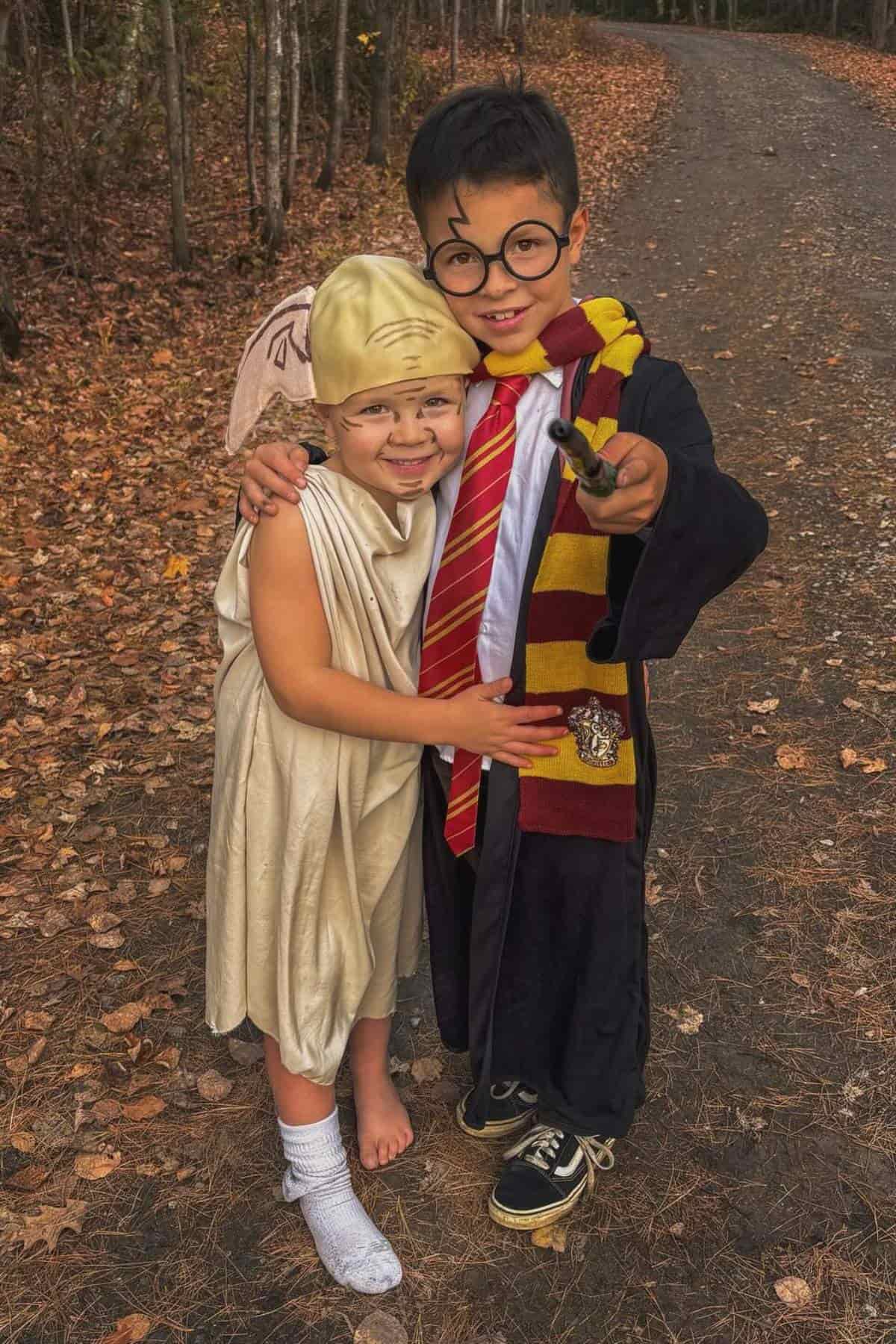 Two children in costumes stand on a leaf-covered path; one is dressed as Dobby the house-elf and the other as Harry Potter, holding a wand and wearing glasses and a Gryffindor scarf.