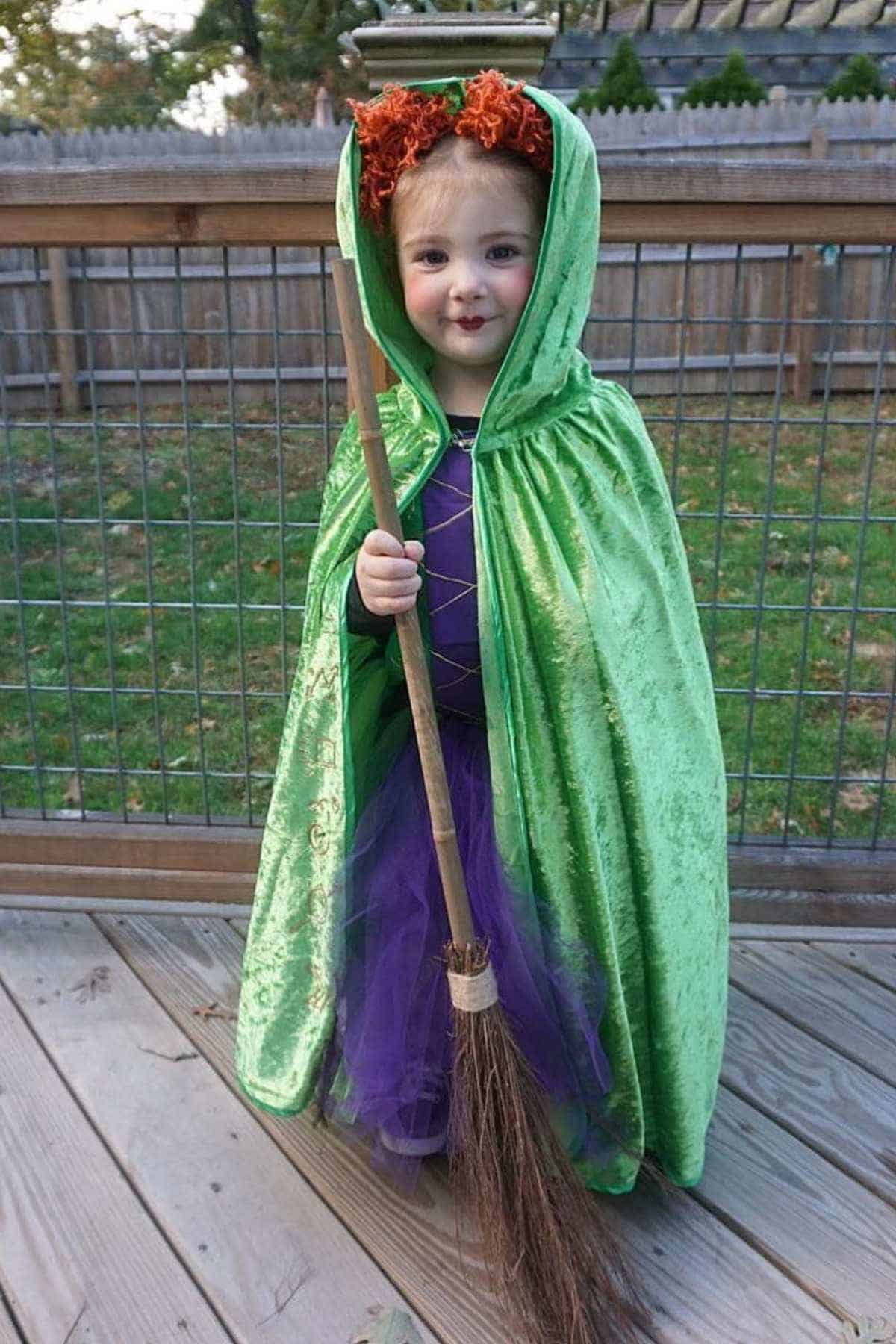 A young child in a green hooded cape and purple dress holds a broom while standing on a wooden deck near a wire fence.