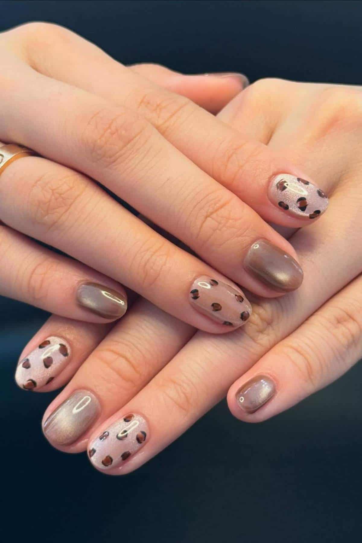 Close-up of hands with short, rounded nails featuring a mix of brown metallic polish and pink nails with black and brown abstract spots.