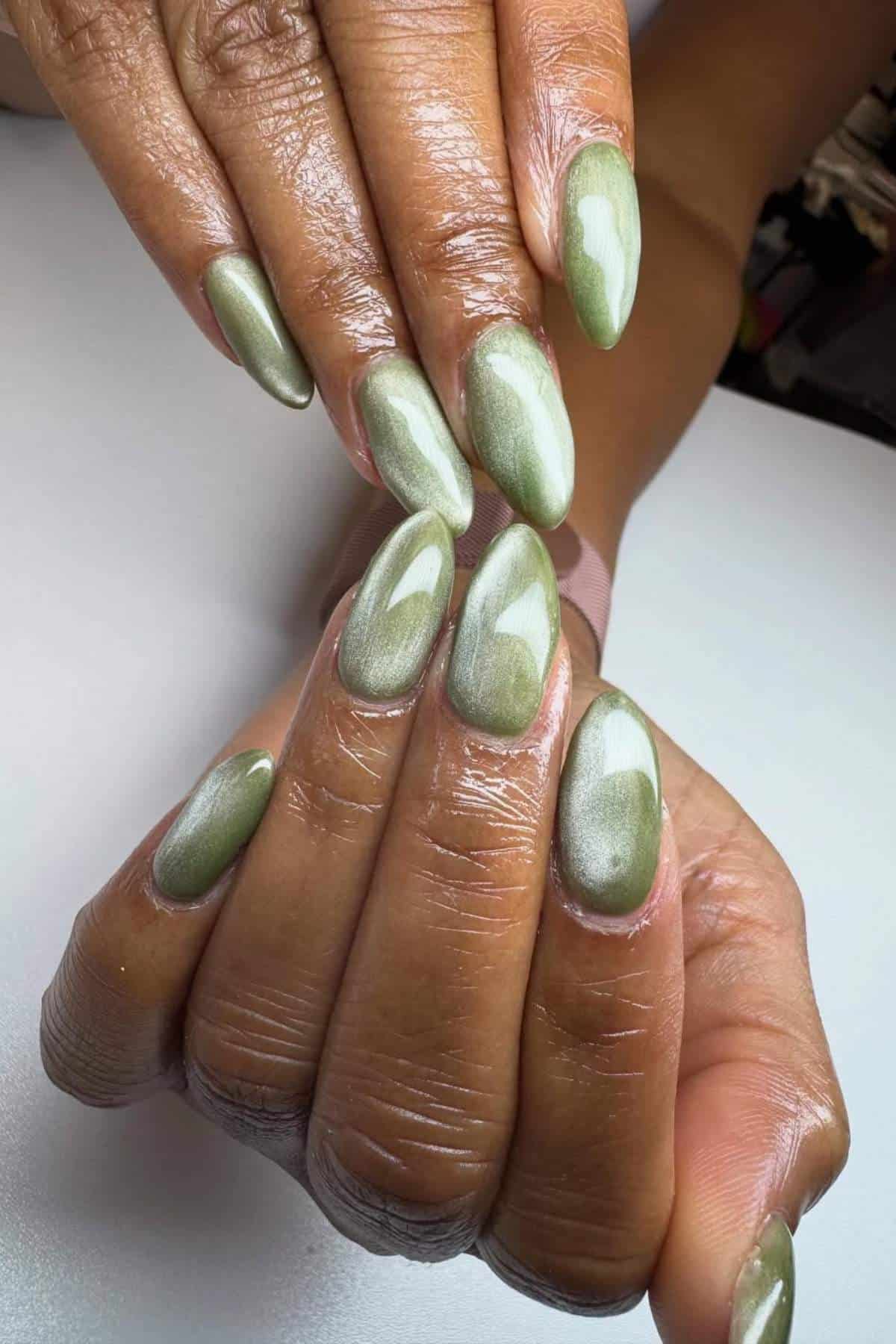 A person displays freshly manicured hands with medium-length, almond-shaped nails painted metallic sage green.