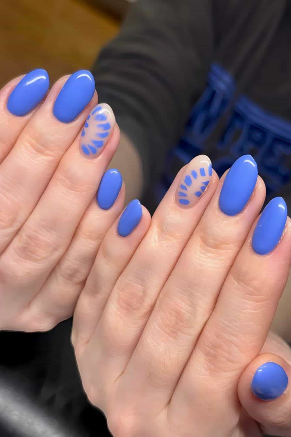 Close-up of hands with oval-shaped nails painted blue, with accent nails featuring a clear base and blue abstract leaf-like designs.
