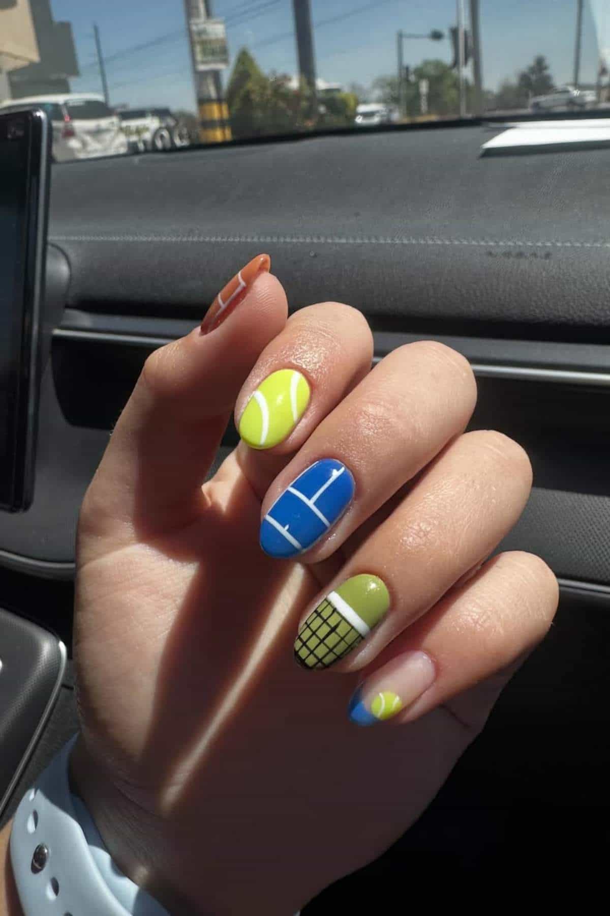 A hand with nails painted in tennis-themed designs, featuring tennis balls, a court, and a net, shown inside a car with sunlight illuminating the nails.