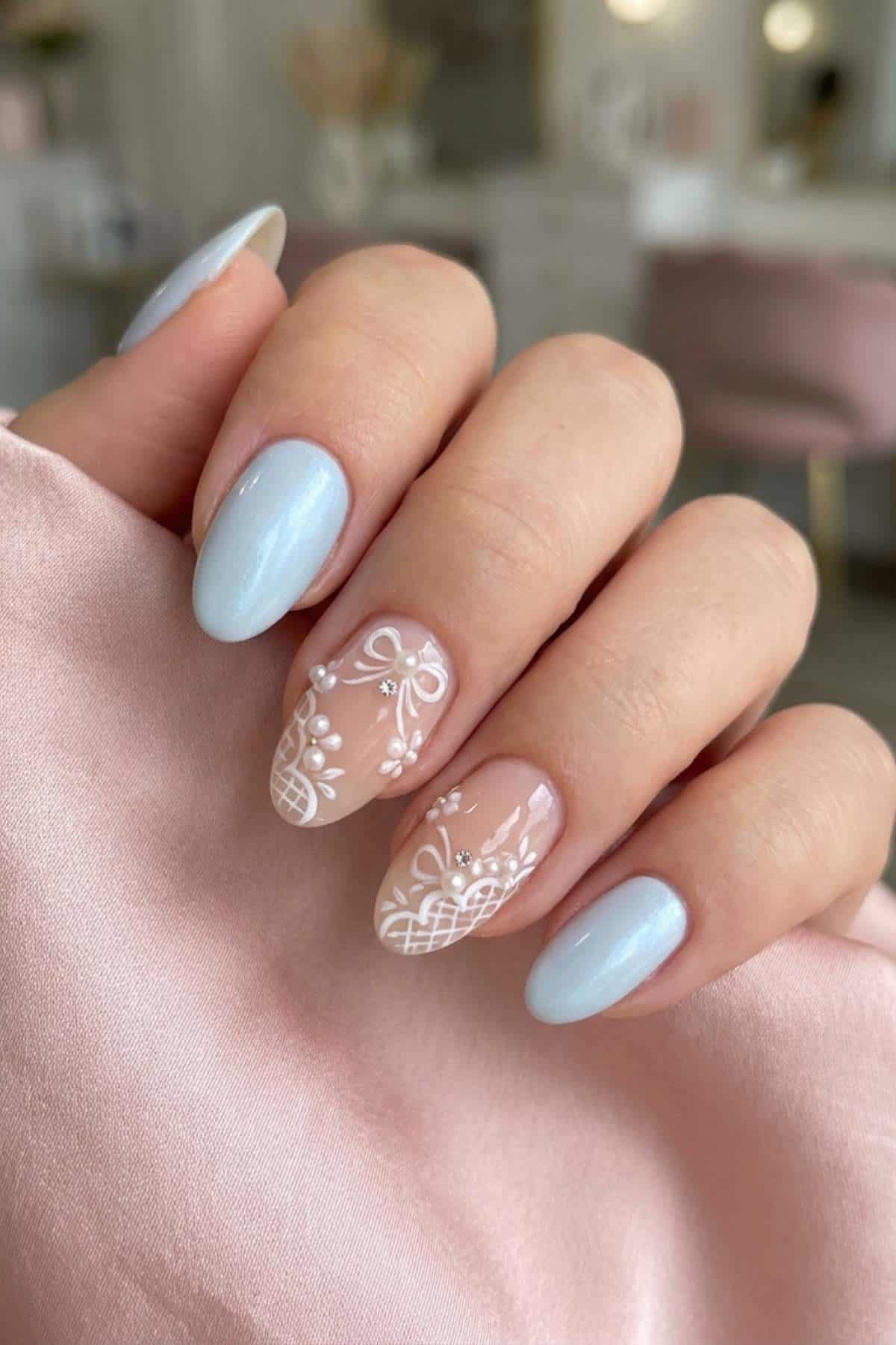 A hand with manicured nails; three nails have pastel blue polish, two feature nude polish with intricate white floral and lace designs.