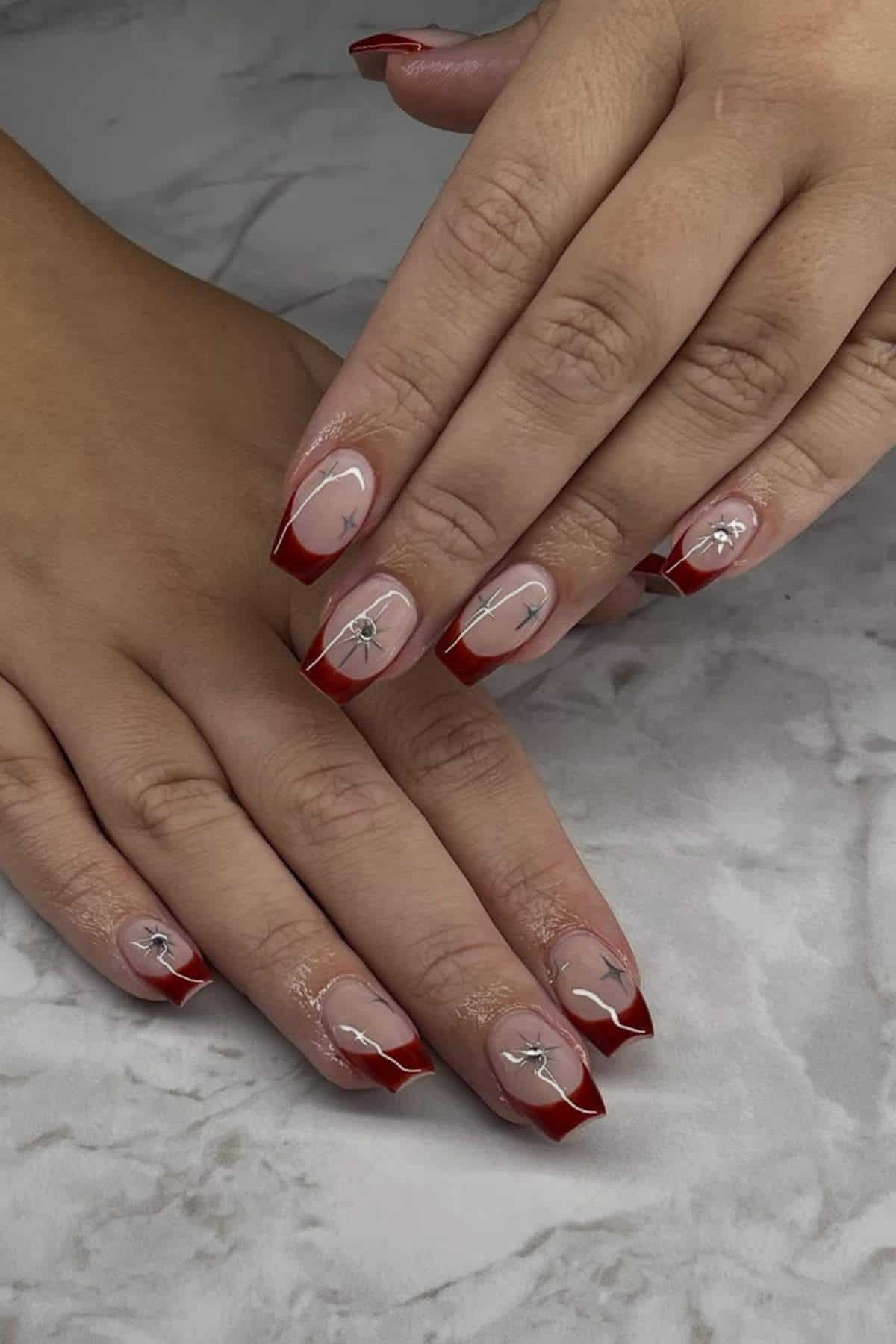 Hands with long ballerina-shaped nails featuring a red French tip and silver bow nail art design on a light marble surface.
