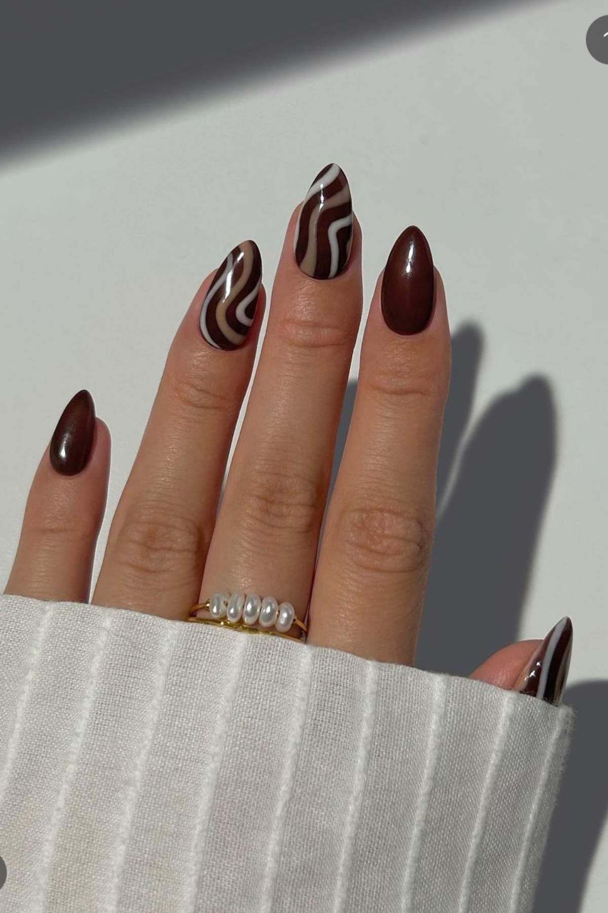 A hand with long, almond-shaped nails painted in glossy brown and beige swirl designs, wearing a gold ring with pearls, rests on a white ribbed fabric background.