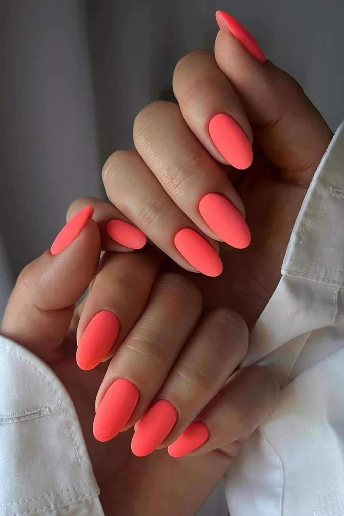 Close-up of hands with long, almond-shaped nails painted in matte neon coral polish, against a blurred neutral background.