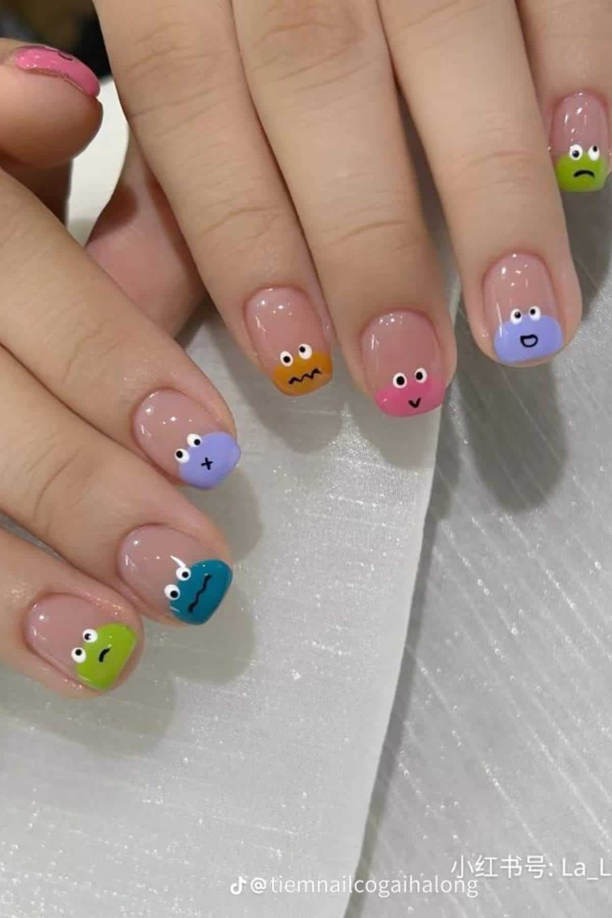 Fingernails painted with pastel tips, each featuring a different cartoon face with various expressions.