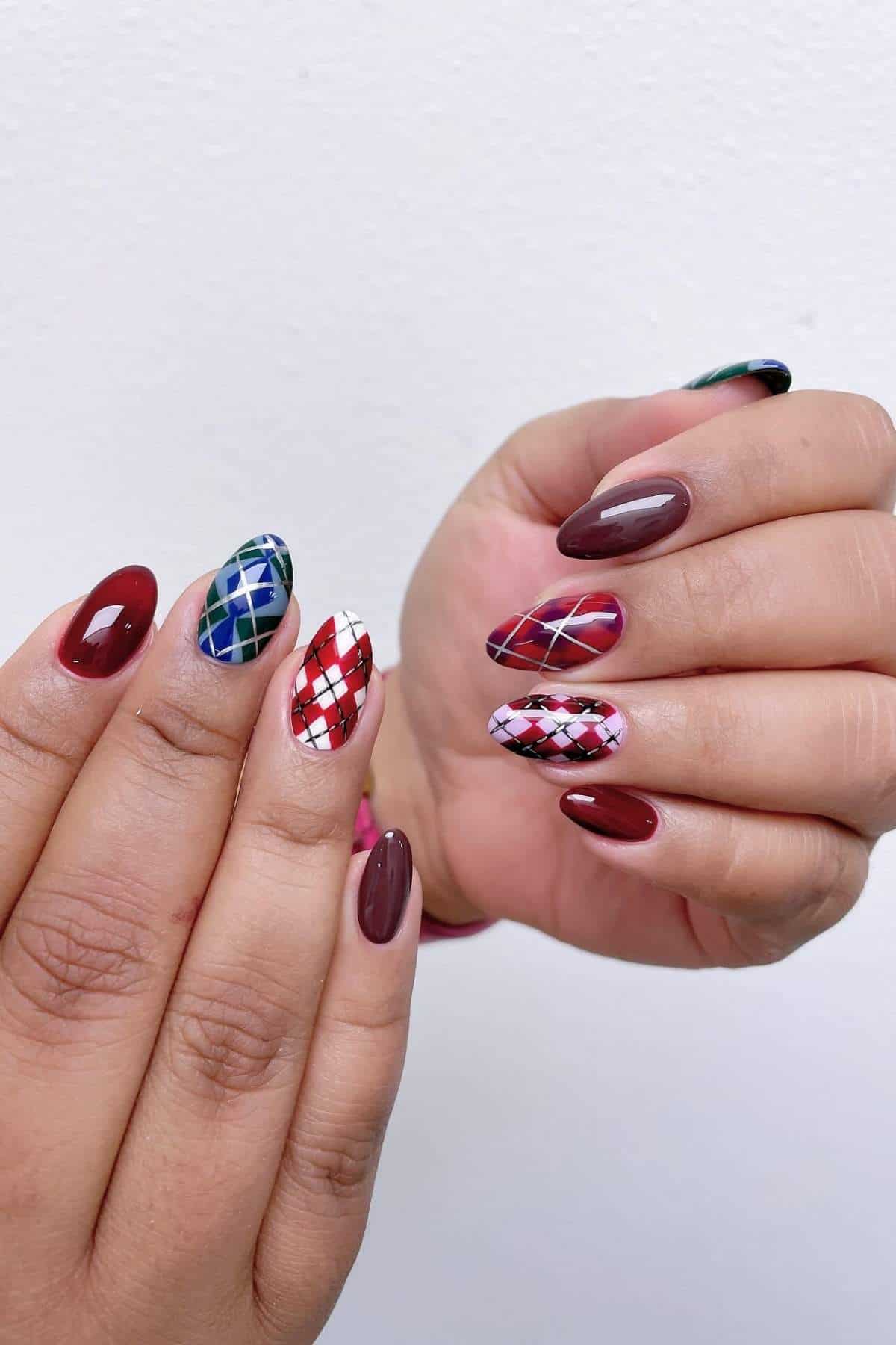 Hands with oval-shaped nails featuring maroon, red, and green plaid designs along with solid maroon and pink accents against a plain white background.