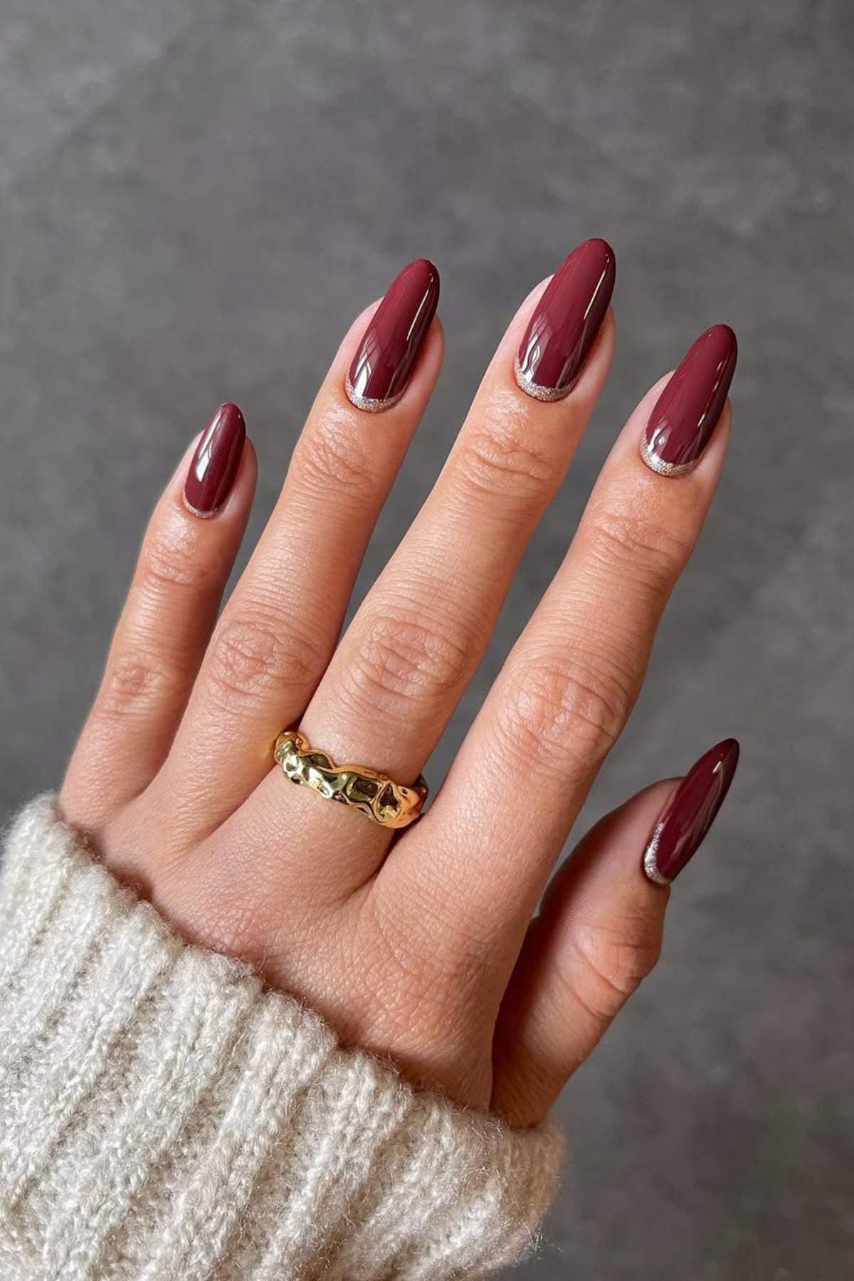 A hand with almond-shaped nails painted in glossy burgundy polish, wearing a gold ring, against a plain gray background.