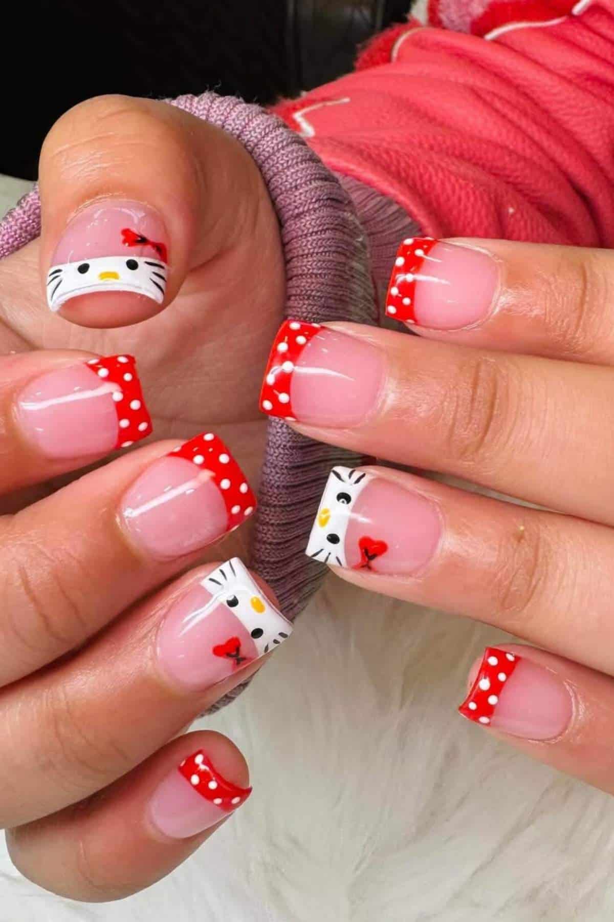 Hands with square-shaped nails featuring red and white polka dot French tips and Hello Kitty designs on some nails. Person is wearing a pink long-sleeve garment.