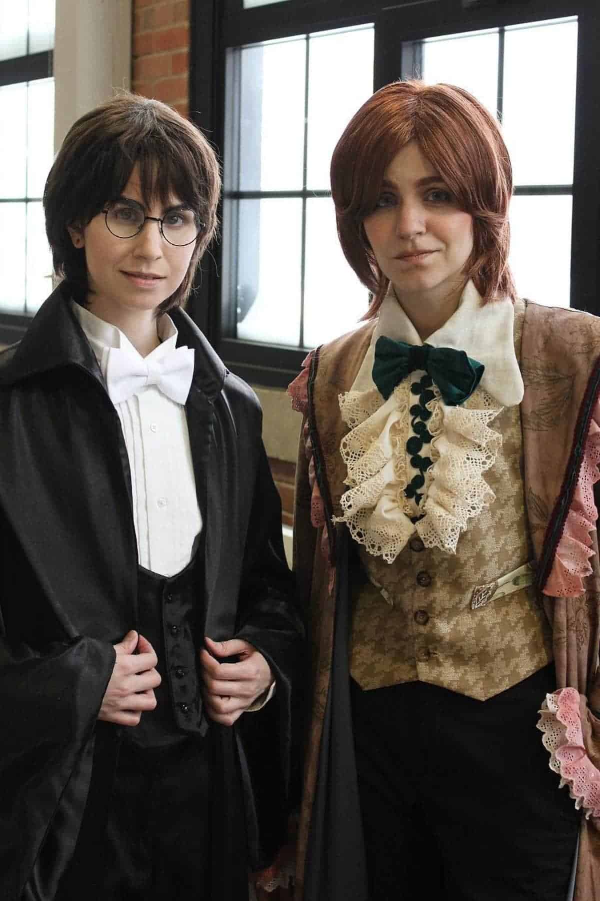 Two people in historical and fantasy-inspired costumes stand indoors; one wears glasses, a bow tie, and a cloak, the other has a lace cravat and a brocade vest.