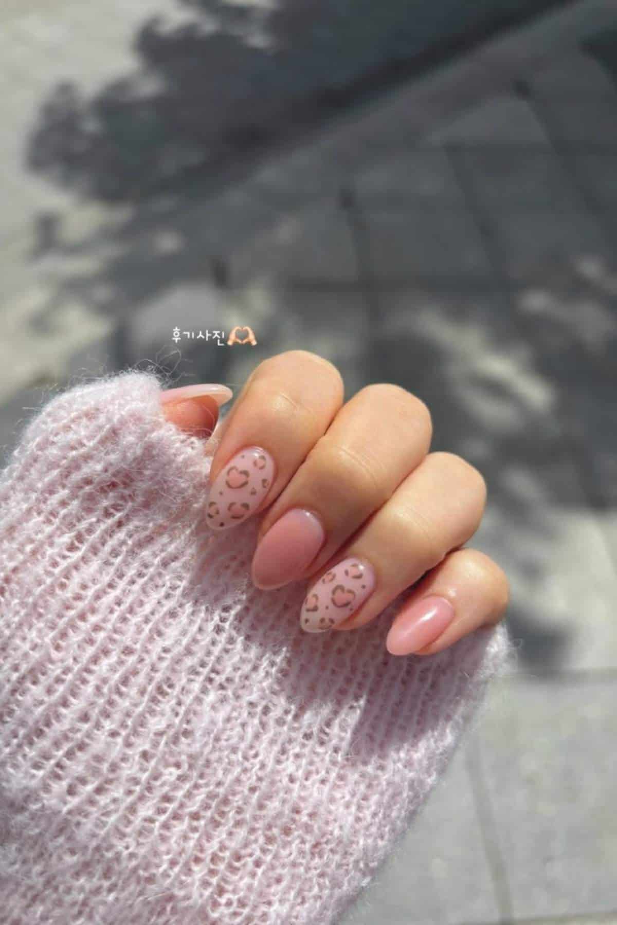 A hand with almond-shaped nails painted light pink, featuring brown leopard print accents, is shown against a soft pink knitted sweater and outdoor background.