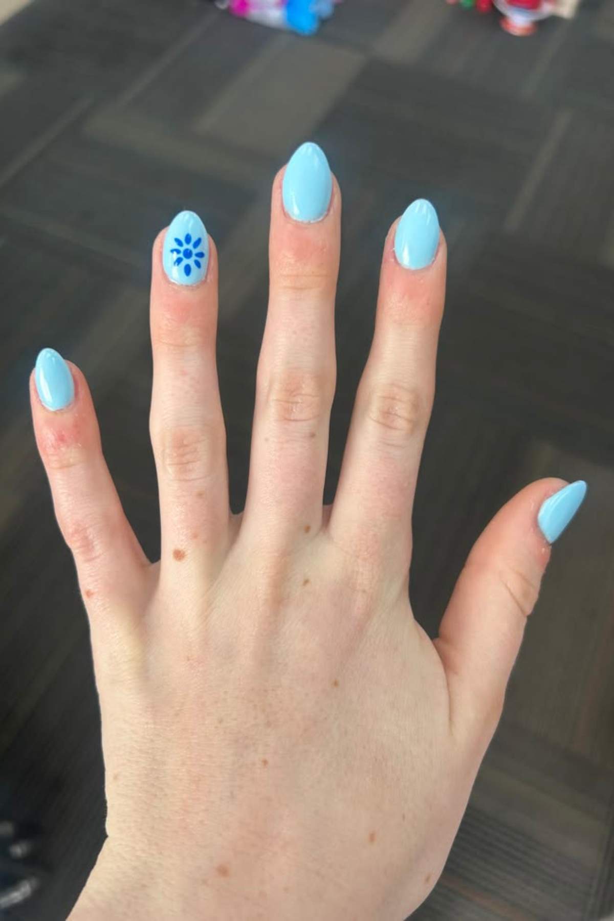 A hand with light blue nail polish, featuring a blue floral design on the ring fingernail.