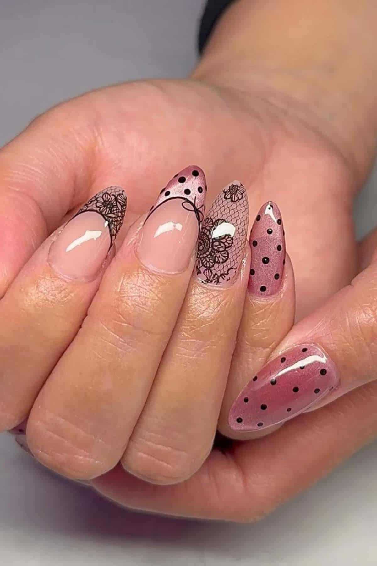 A hand with almond-shaped nails featuring a pink base, black floral designs, and black polka dots on alternating fingers.
