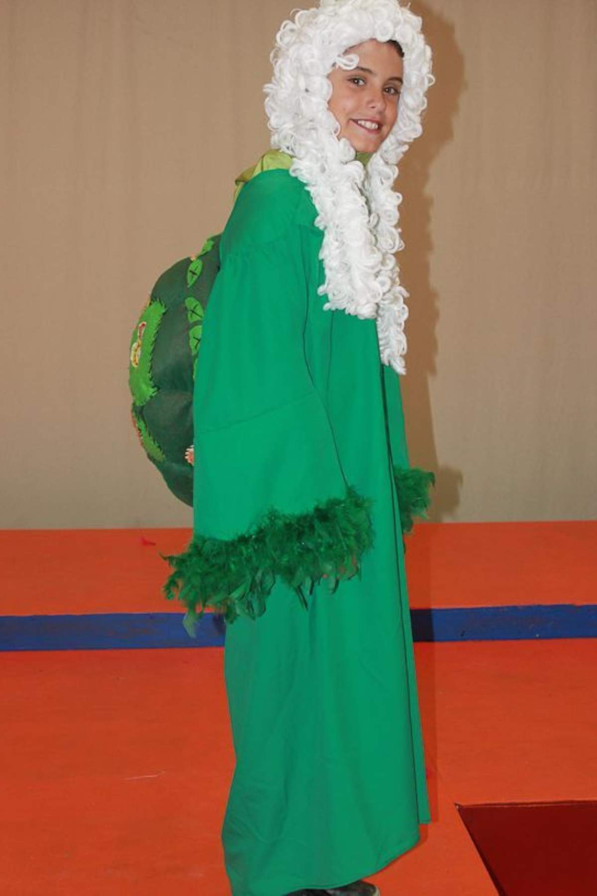 A child wearing a green robe with feathered cuffs, a white curly wig, and a turtle shell prop on their back stands on an orange and blue floor.