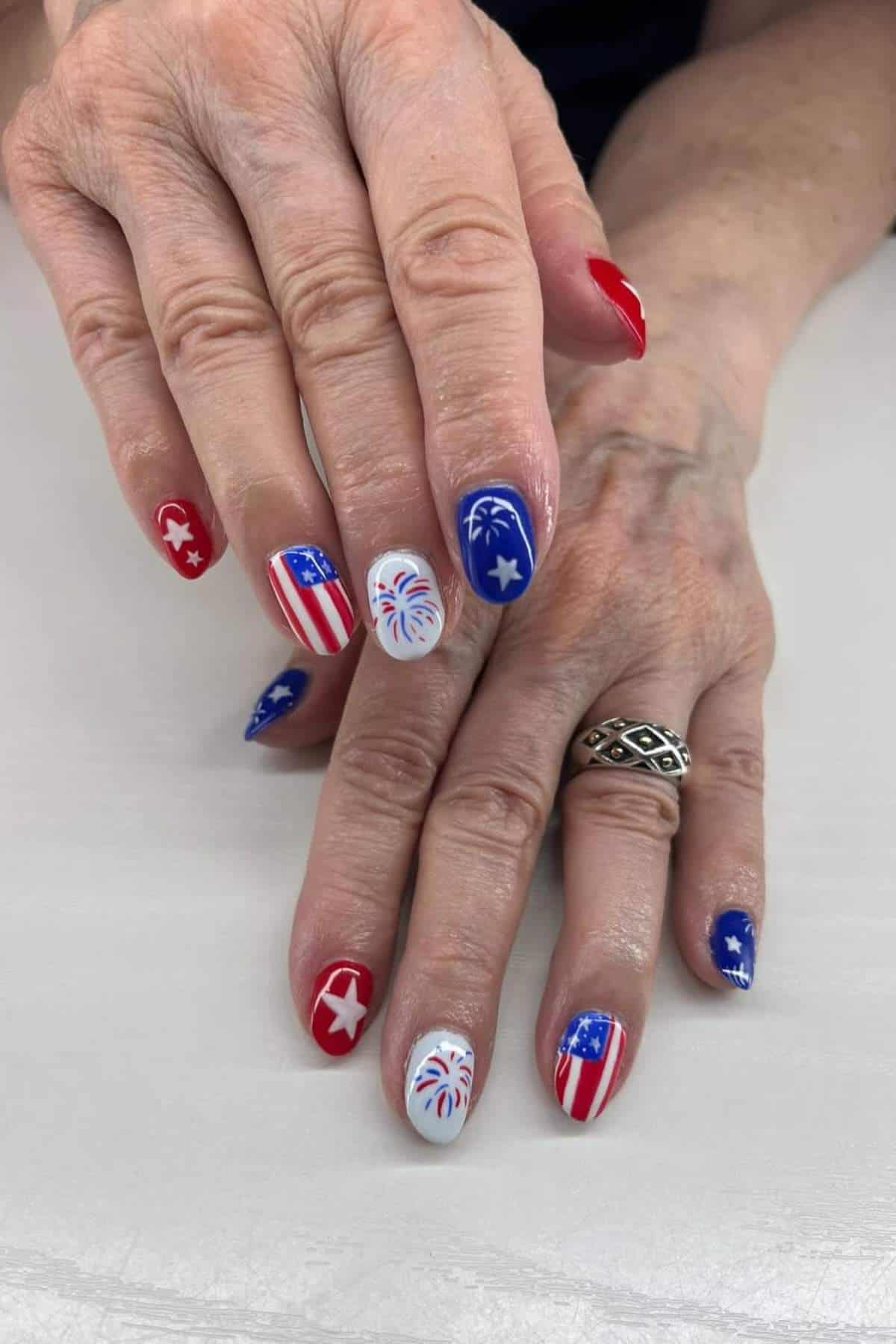 Hands with manicured nails featuring red, white, and blue designs, including stars, stripes, and fireworks. The person wears a patterned silver ring on one finger.